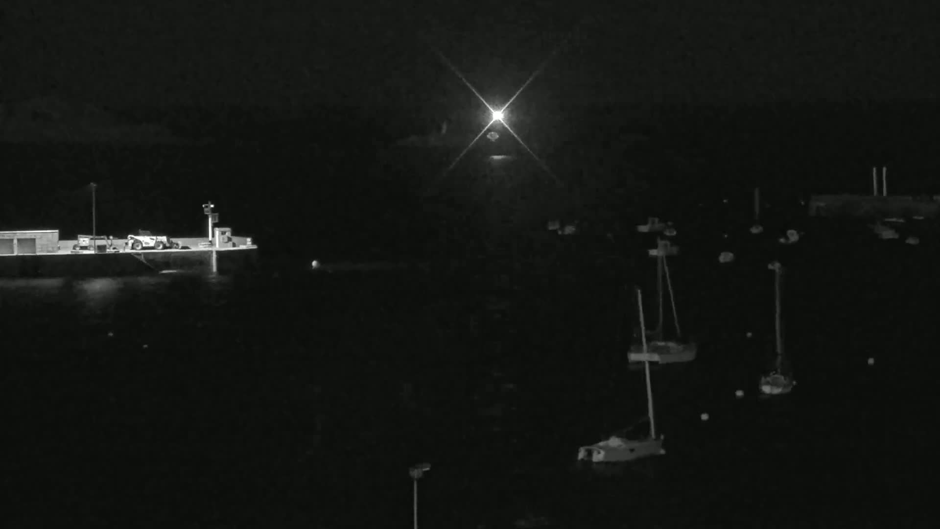 A nighttime harbor scene shows a building with a piece of machinery on a dock, several sailboats moored in the water, and a bright light shining in the distance.