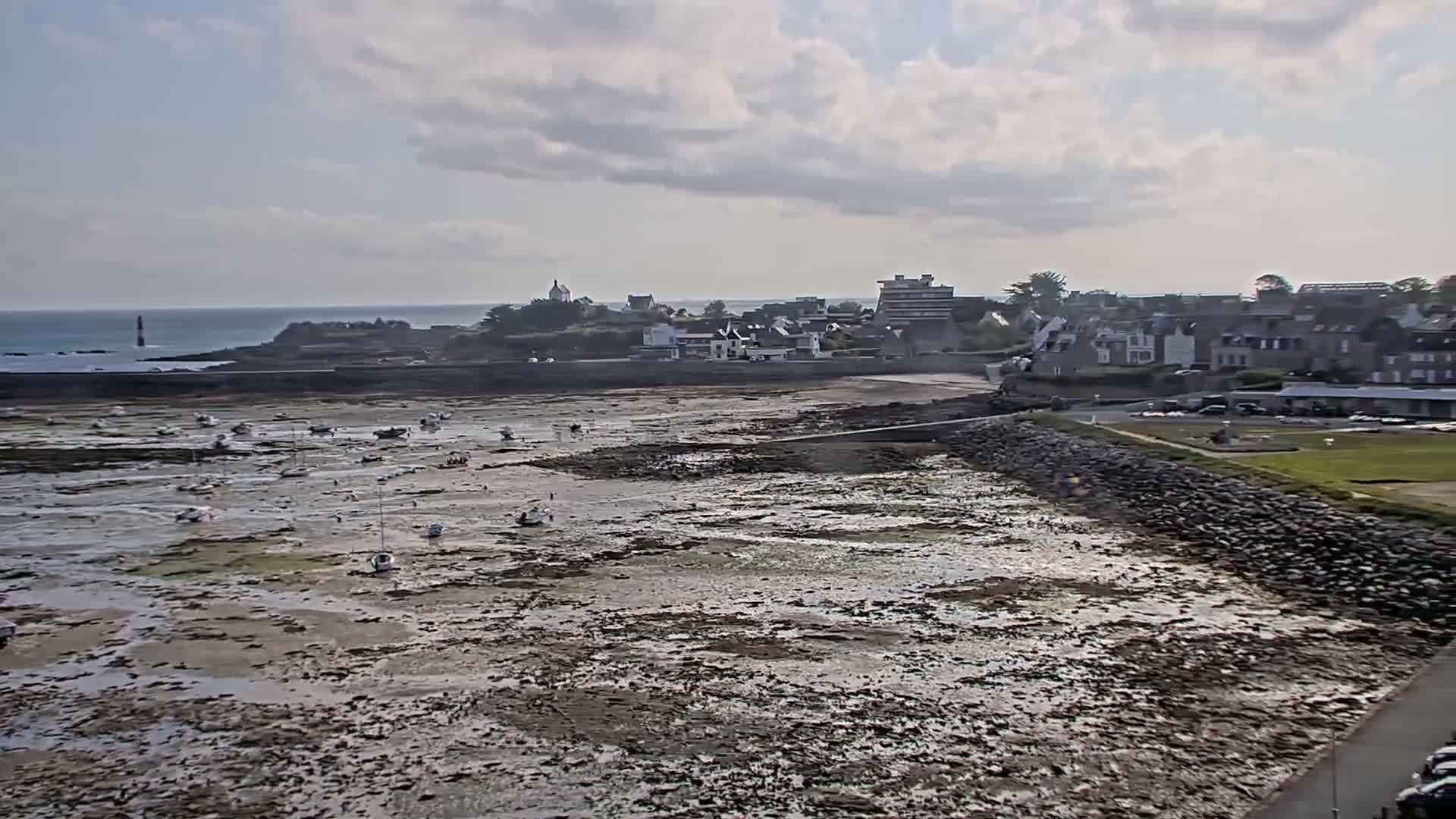 A coastal town is seen under a partly cloudy sky, with numerous boats resting on the exposed seabed at low tide.