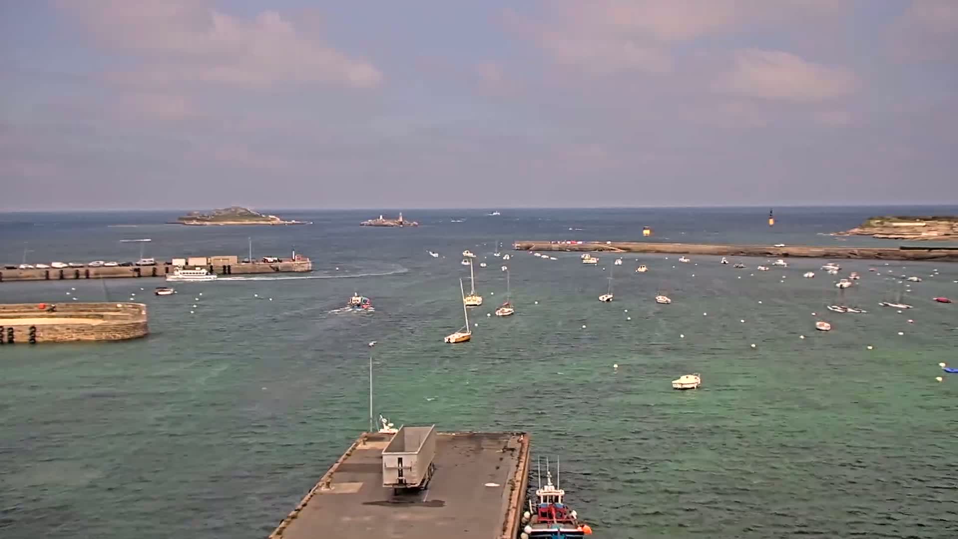 A harbor filled with sailboats and small motorboats under a partly cloudy sky, with a distant island and a larger vessel near the harbor entrance.