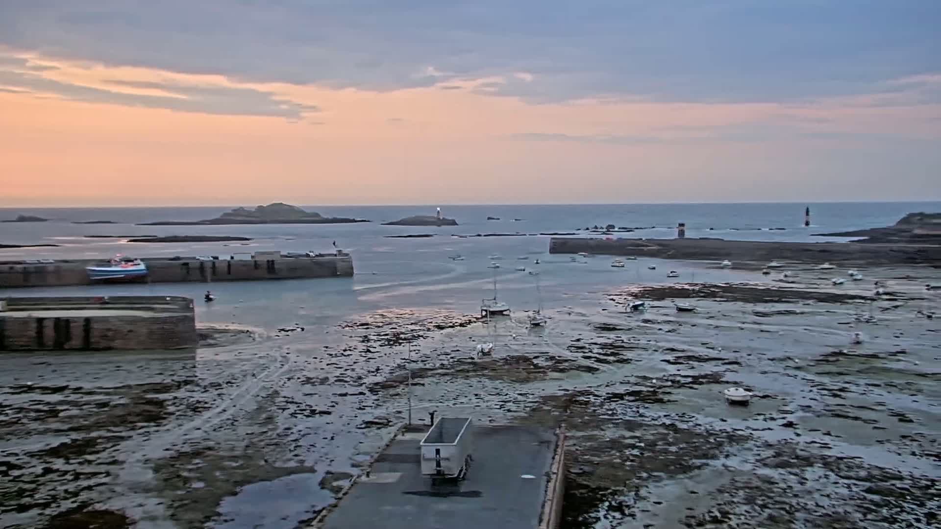 A harbor at low tide during sunset, with numerous small boats grounded on the exposed seabed, and a larger vessel moored inside a stone harbor wall.