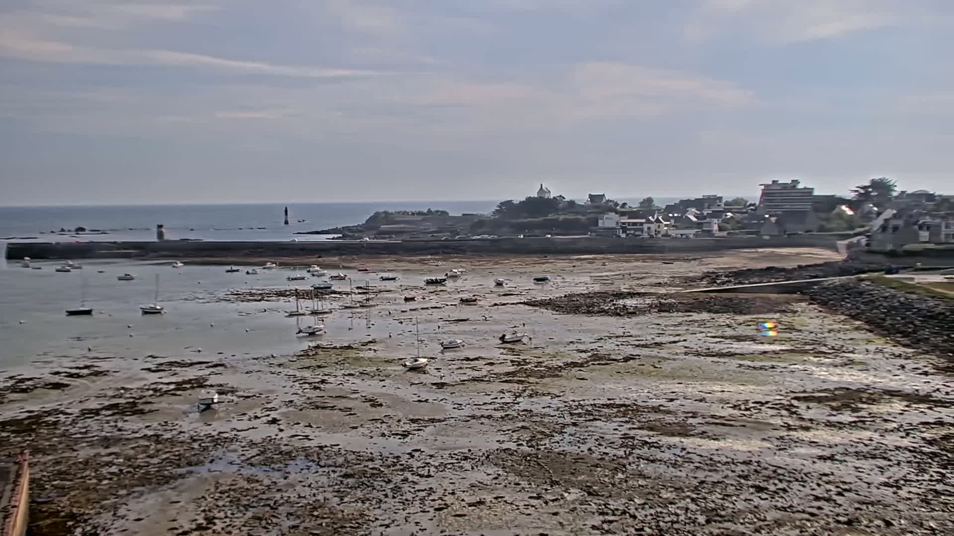 A coastal town is seen across a wide expanse of exposed mudflats at low tide, with numerous small boats scattered throughout the shallow waters and a calm ocean in the background under a mostly cloudy sky.