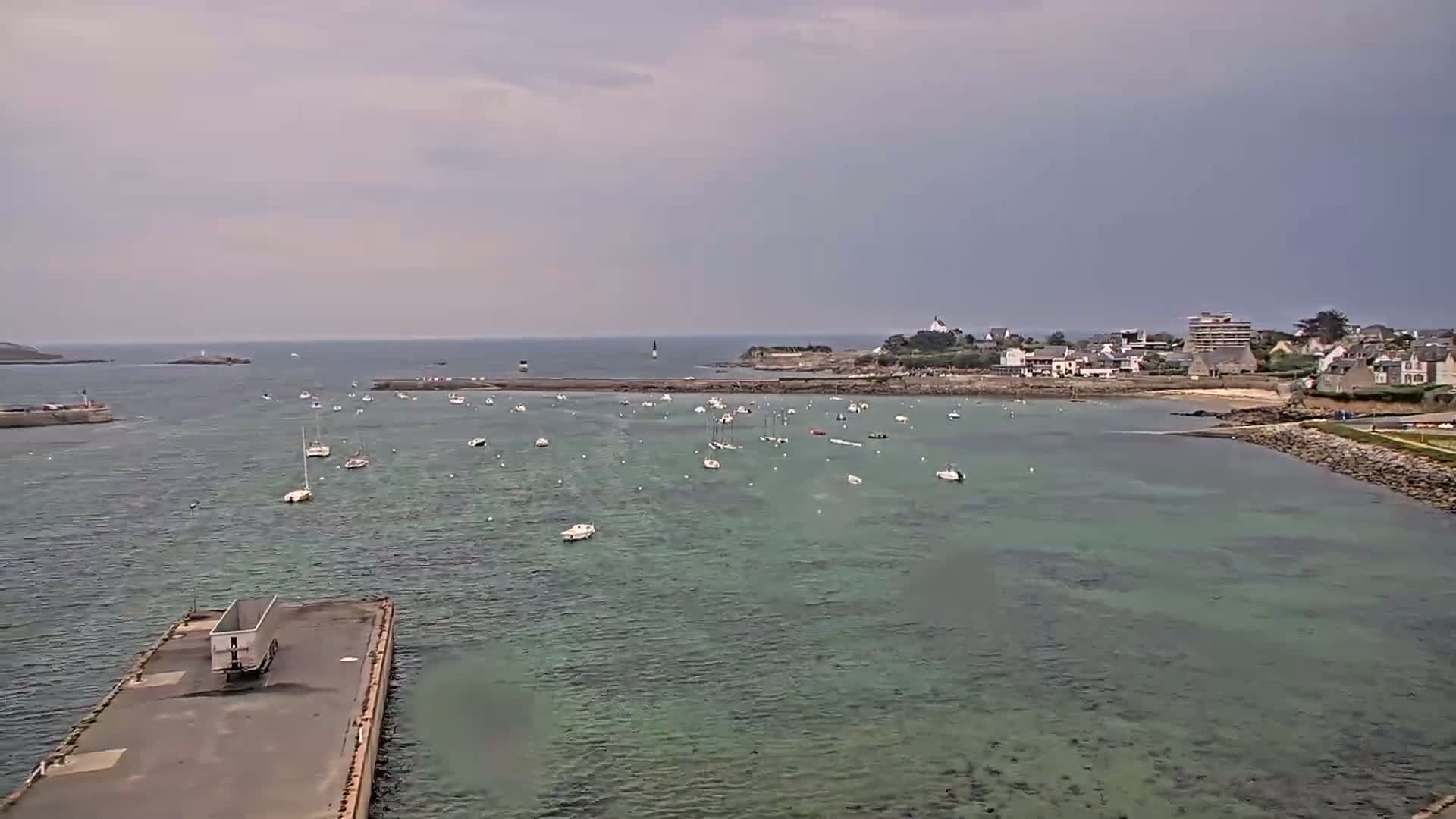 A calm, overcast day shows a harbor filled with numerous small boats, a small town on a shoreline, and a larger body of water extending to the horizon.