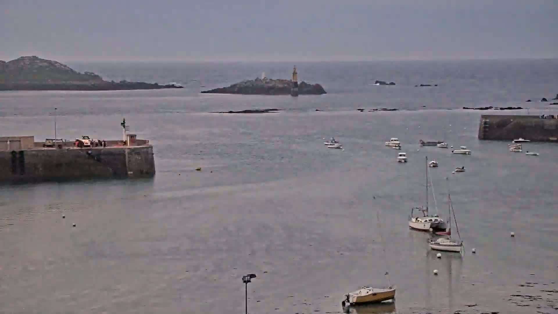 A calm, overcast day shows a harbor with several small boats and a lighthouse on a small island in the distance.