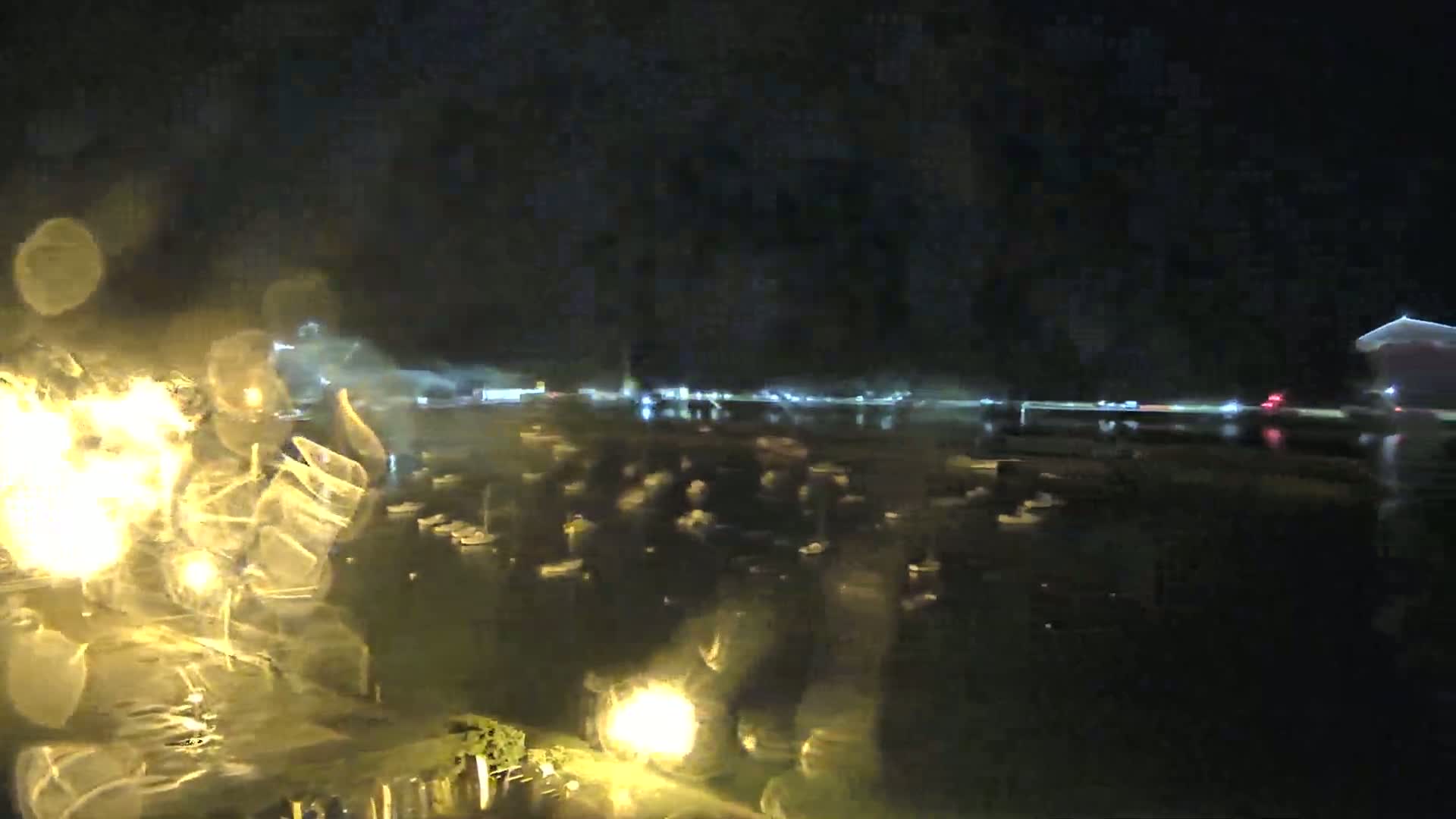 A nighttime view through rain-covered glass shows a harbor filled with many boats, and a distant town lit up with lights.