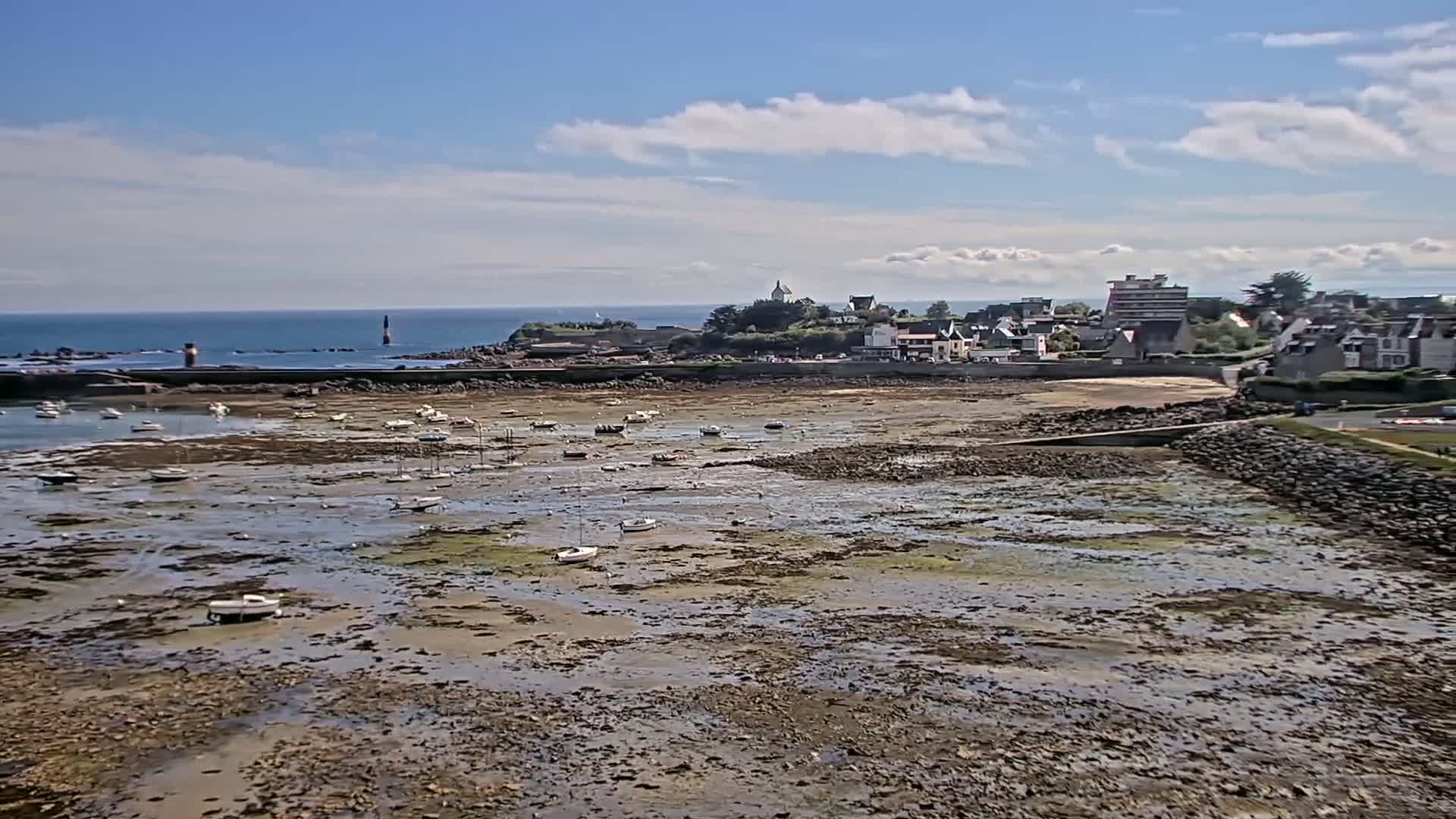 Under a mostly sunny sky, a coastal town overlooks a harbor with numerous small boats resting on exposed mudflats at low tide.