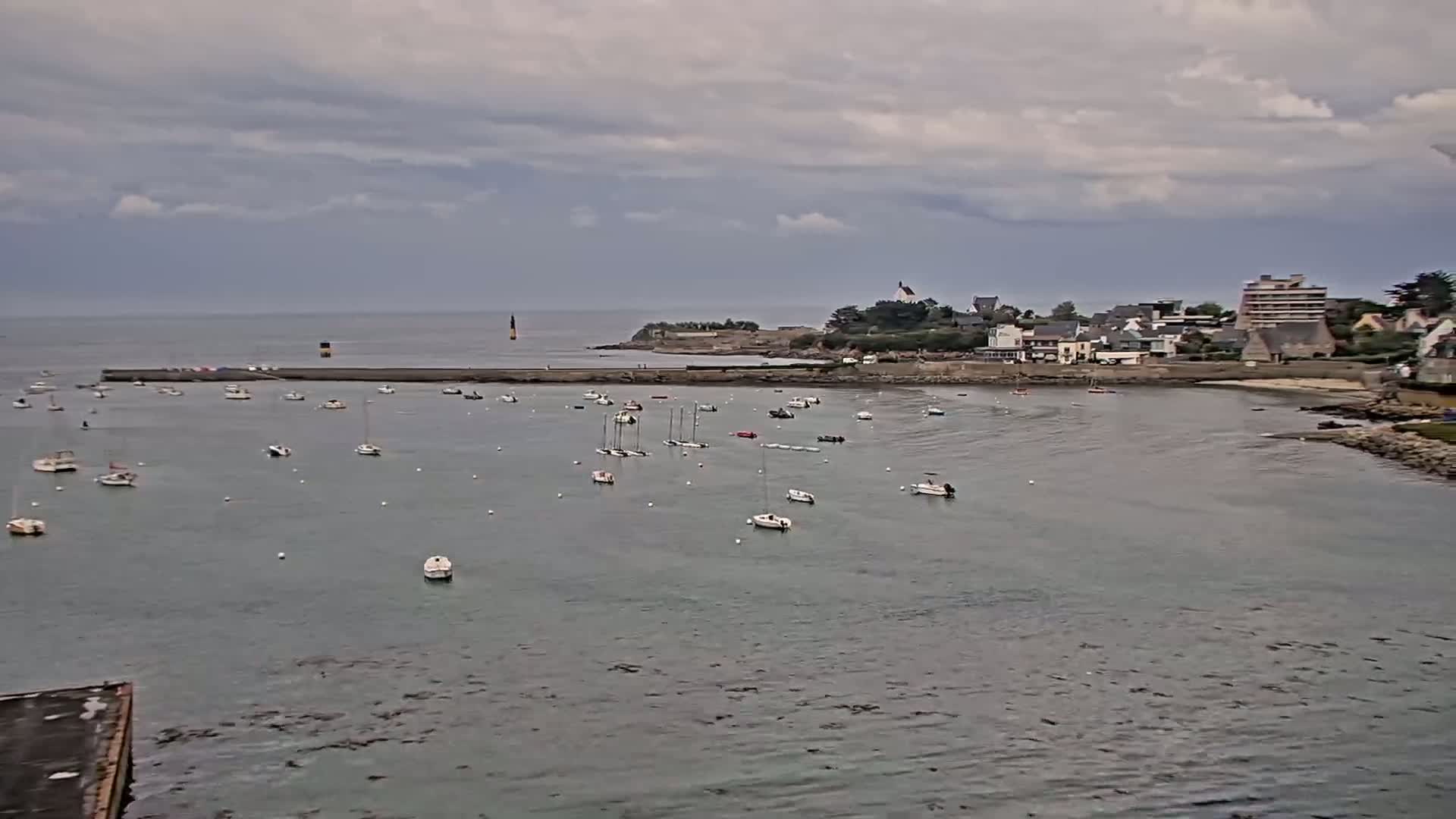 Numerous sailboats are moored in a calm bay under a cloudy sky, near a small town situated on a peninsula.