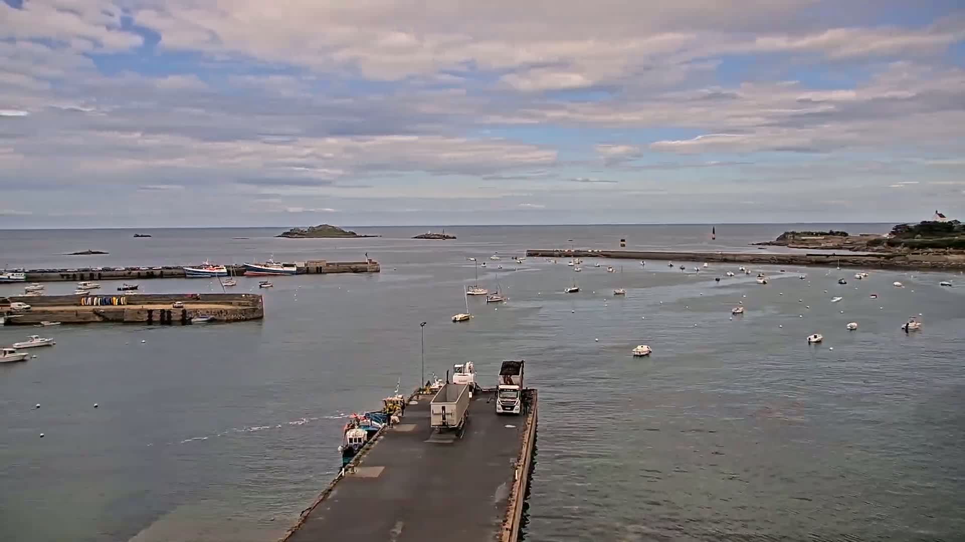 A harbor filled with numerous boats under a partly cloudy sky, with a pair of trucks parked on a pier extending into the water.