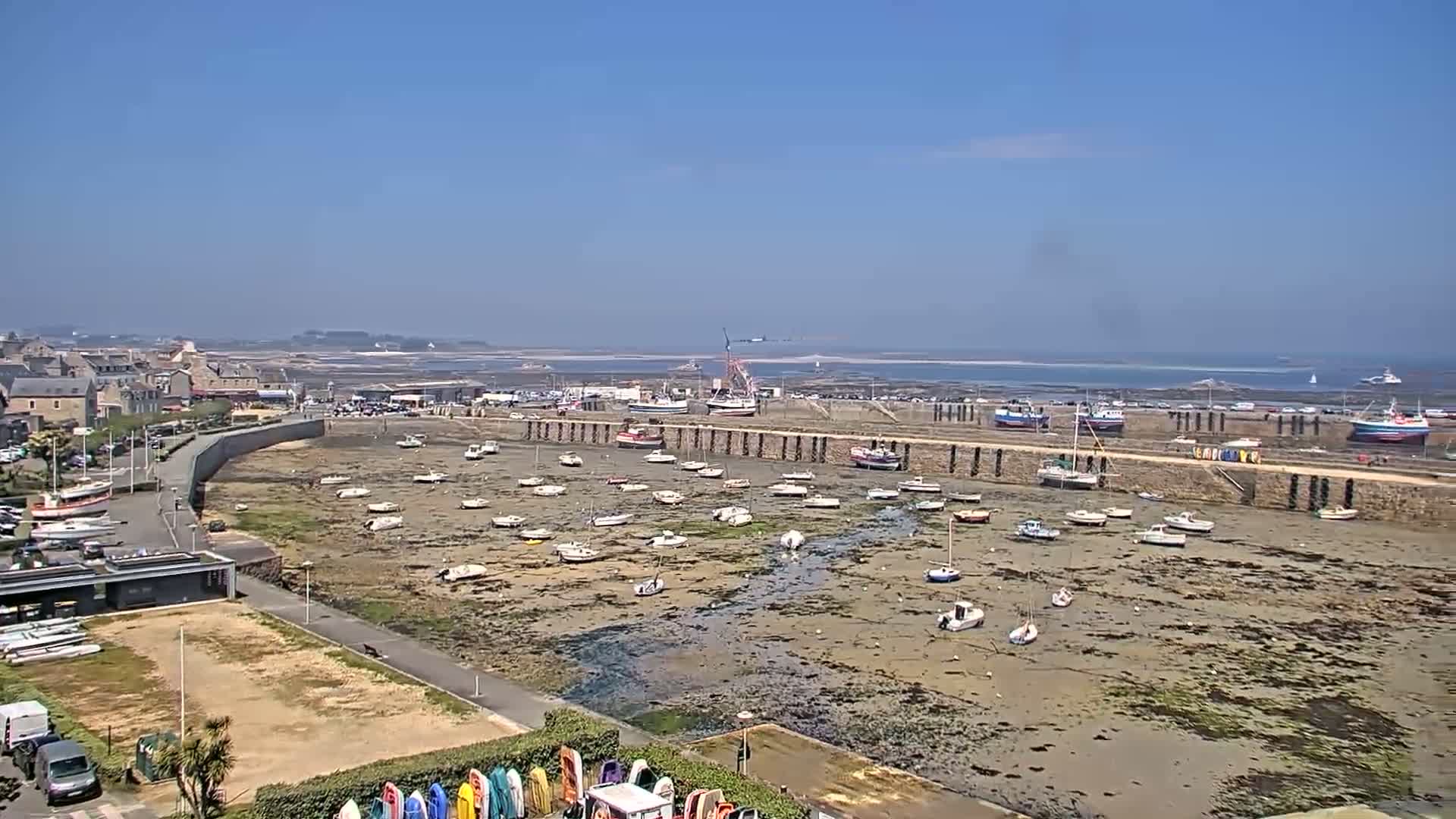 A harbor filled with numerous small boats sits exposed at low tide on a sunny day, with a town and ocean visible in the background.