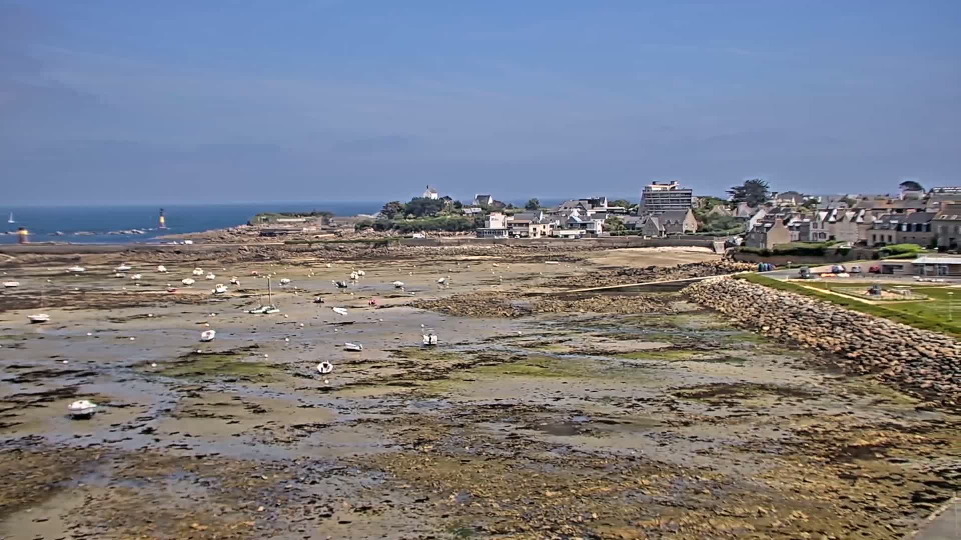 A coastal town with numerous small boats sits on a rocky shoreline under a clear blue sky, with the tide out, exposing a vast expanse of mudflats.
