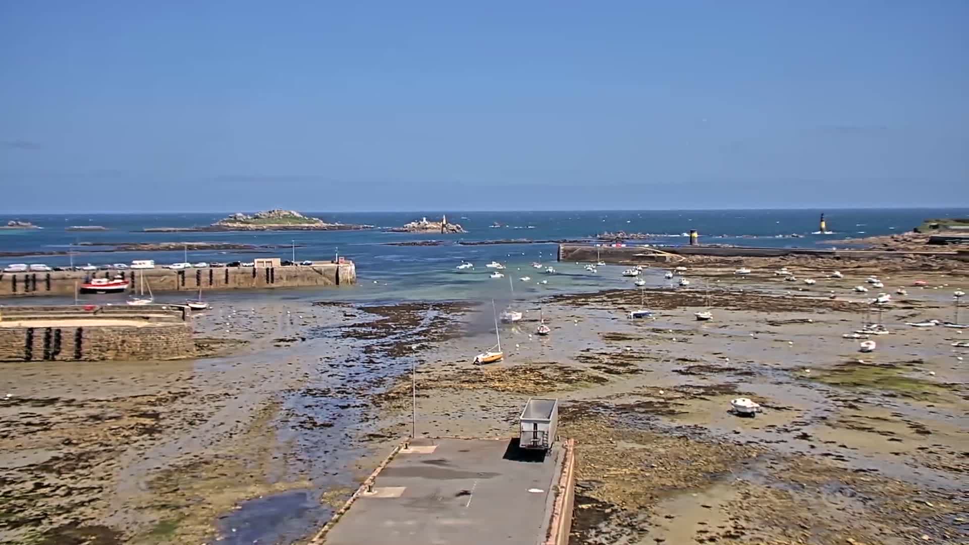 A harbor with numerous small boats sits mostly exposed at low tide on a sunny day, with islands and a lighthouse visible in the distance.
