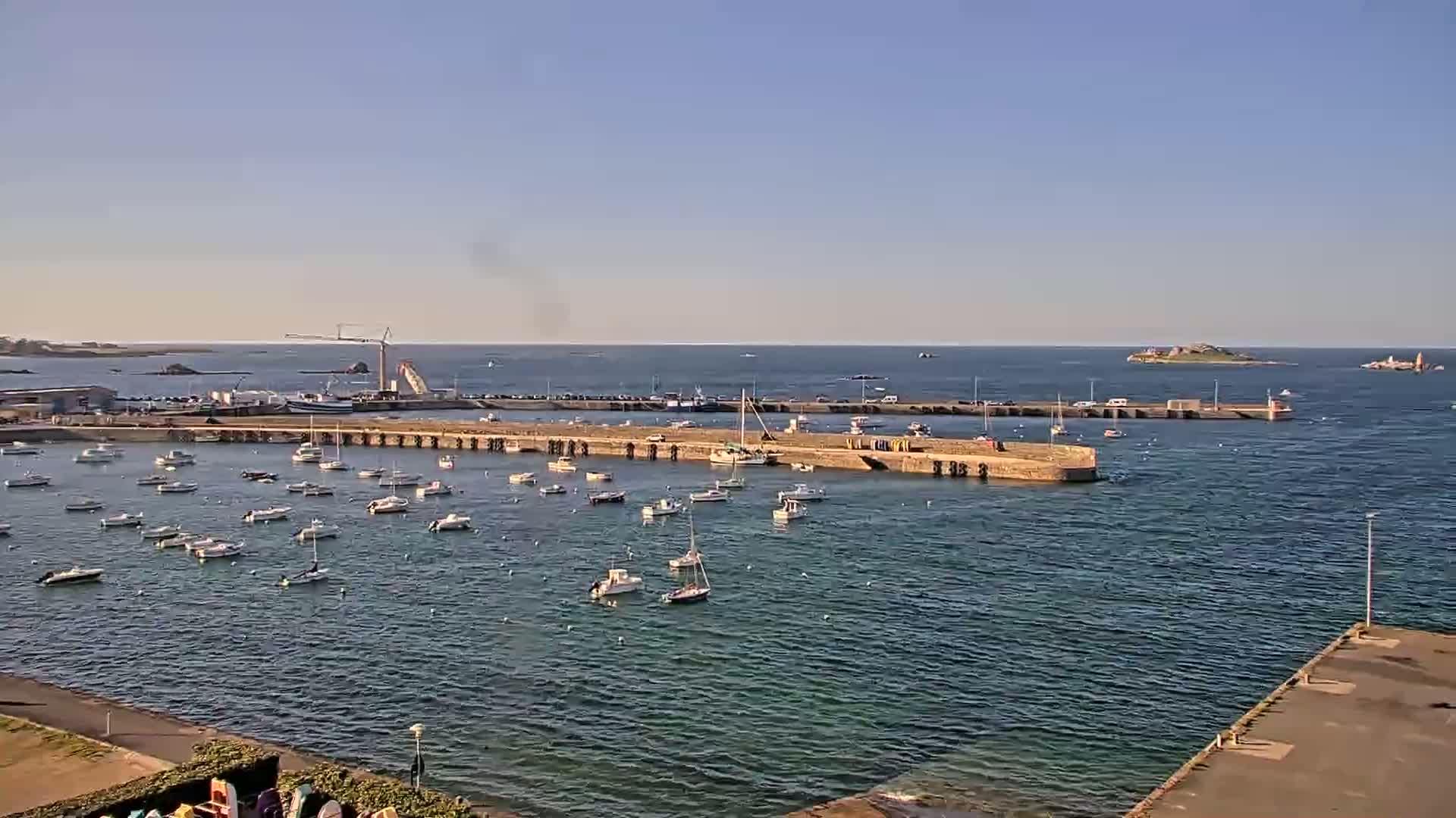 A harbor filled with numerous small boats under a clear, sunny sky, with a larger body of water and distant islands visible in the background.
