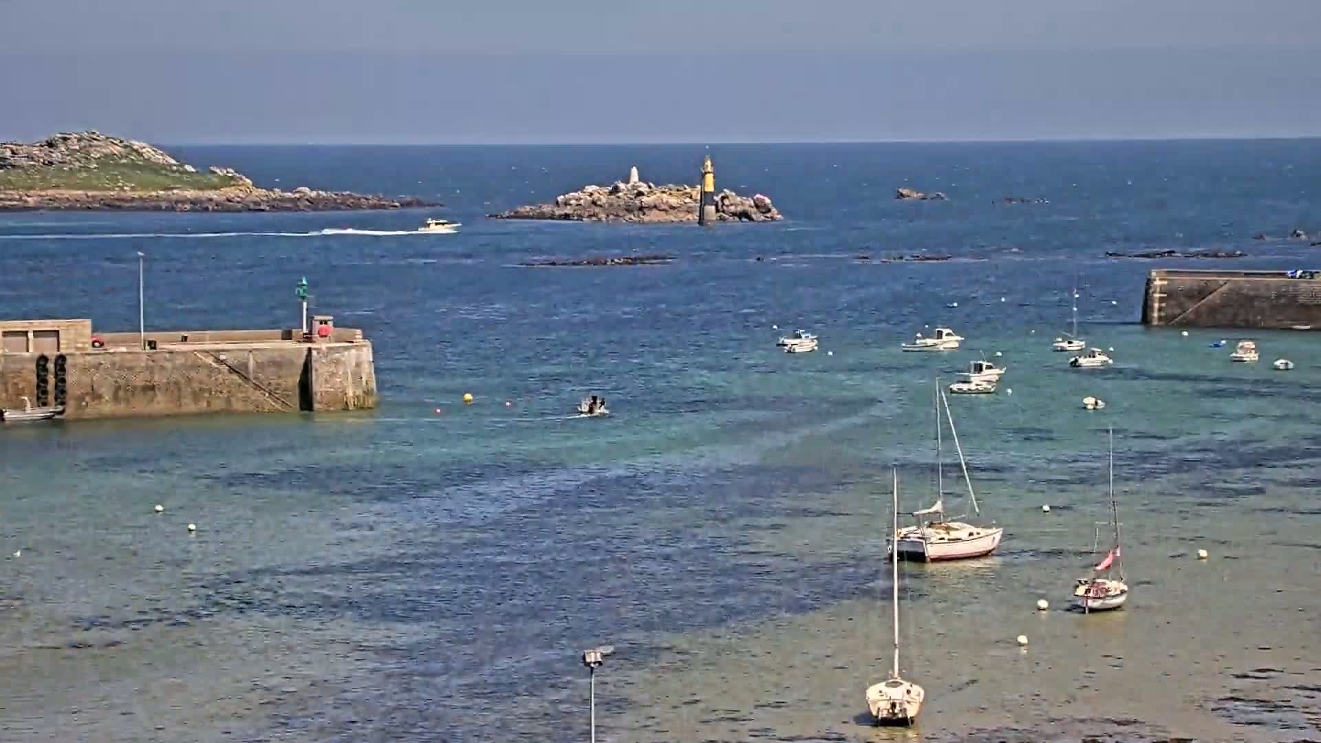 A calm, sunny day shows a harbor with several sailboats and motorboats moored near stone piers and a small island with a lighthouse in the distance.