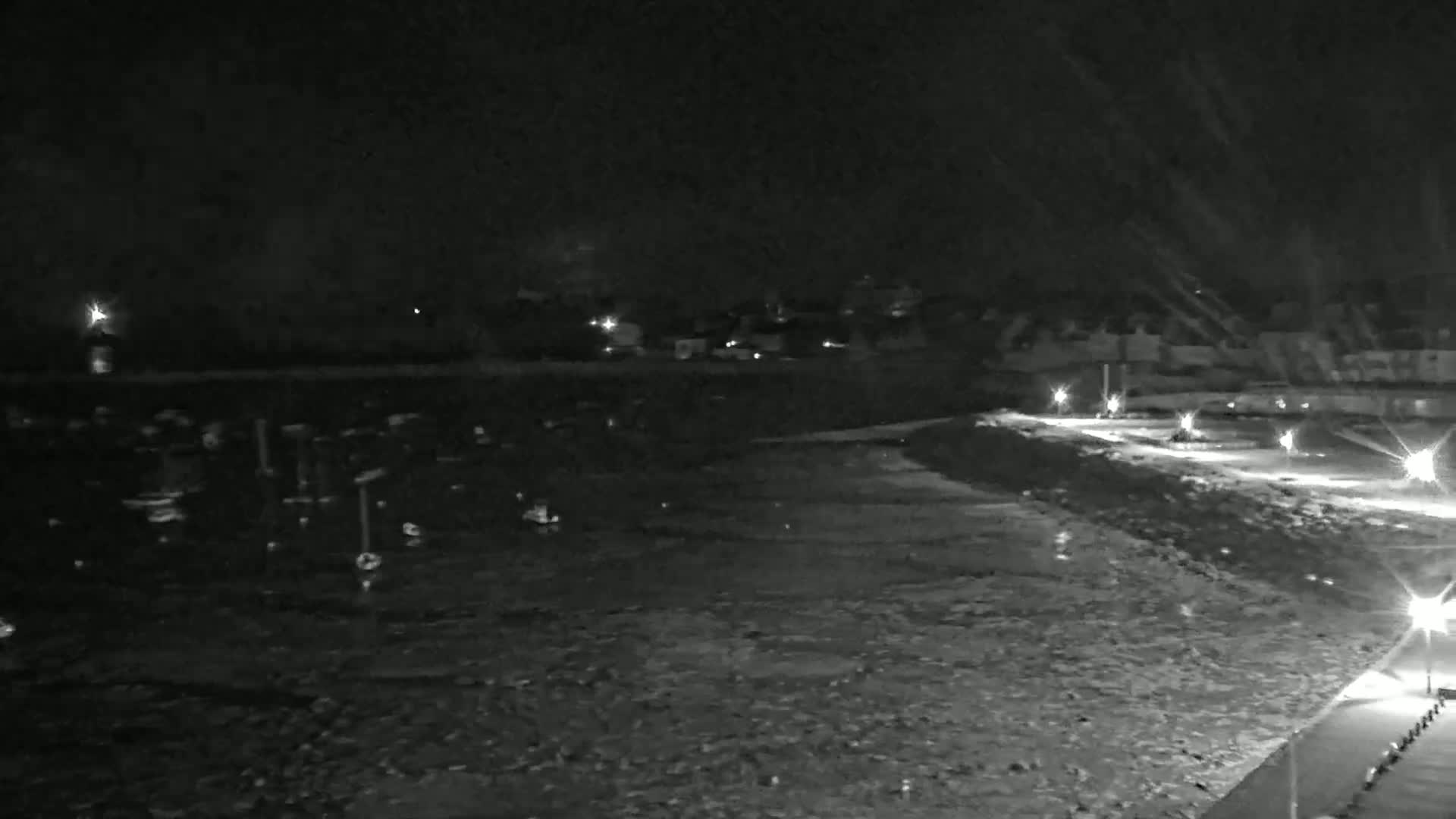A nighttime, snow-covered waterfront scene shows a dark body of water with faintly visible objects, a lit walkway, and a dimly lit shoreline with buildings in the background.