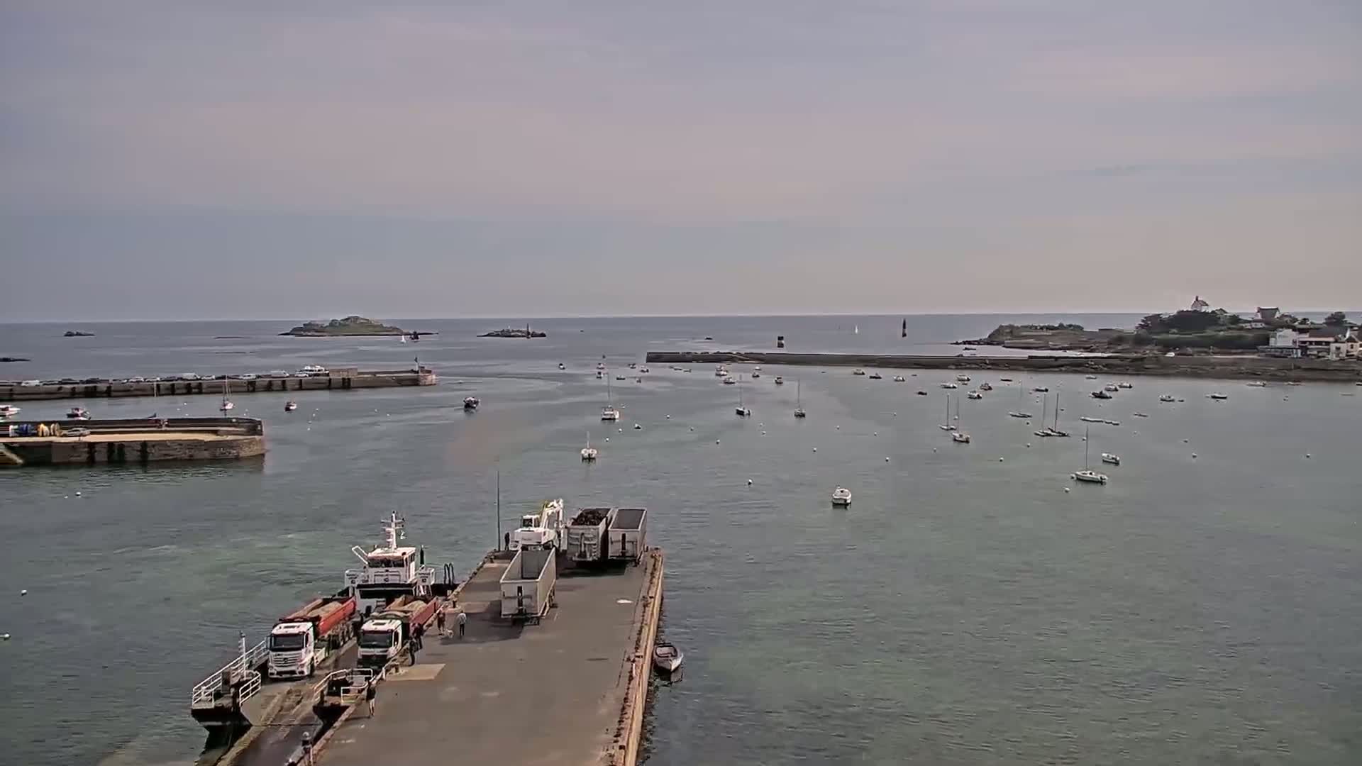 A harbor with numerous boats, a ferry with trucks, and distant islands under a cloudy sky.