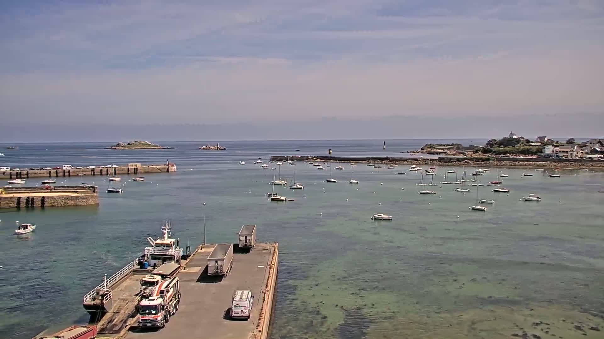 A harbor filled with numerous sailboats and small motorboats under a partly cloudy sky, with a ferry docked and trucks parked on a pier.