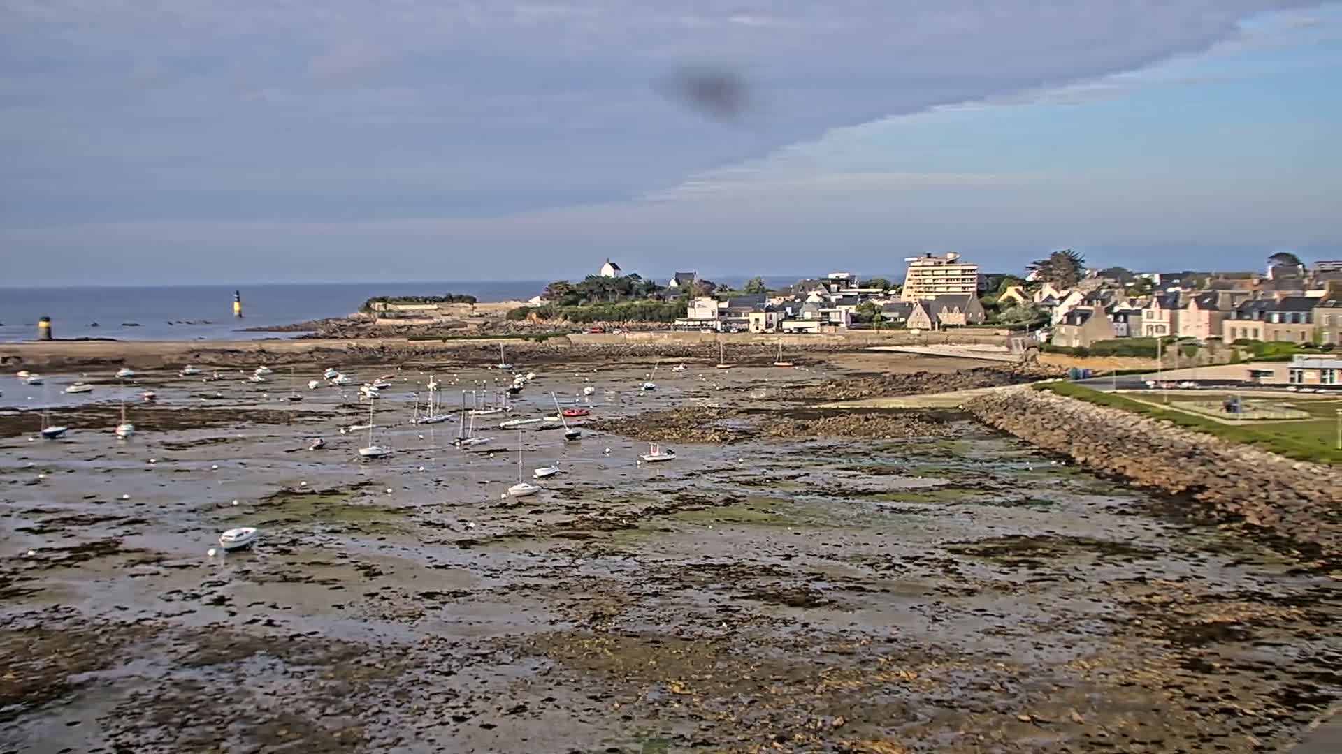 A mostly cloudy day reveals a coastal town with numerous sailboats resting in a harbor at low tide.