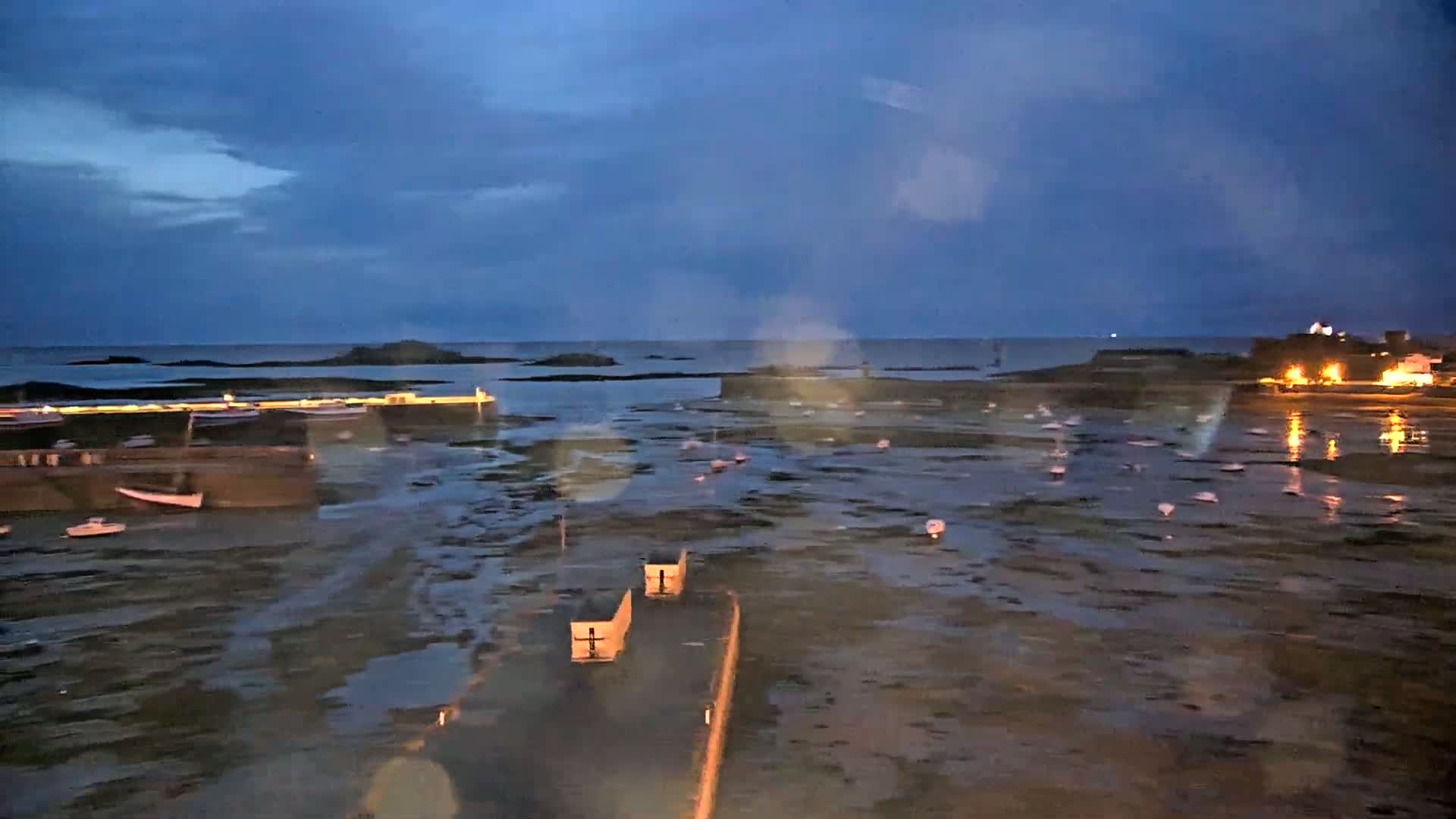 A low-light view of a harbor at low tide, with boats scattered across the exposed seabed under a dark blue, possibly rainy sky.