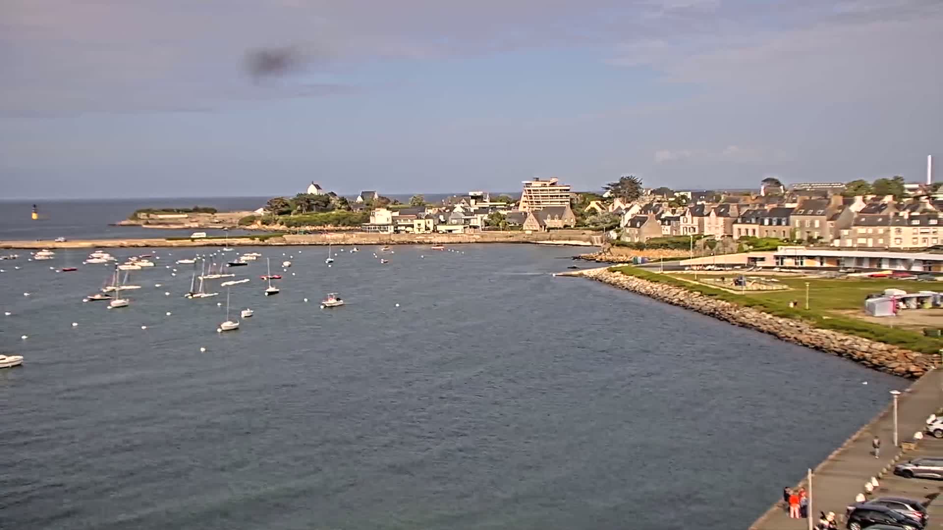 A calm harbor filled with sailboats is situated in front of a small town on a partly cloudy day.