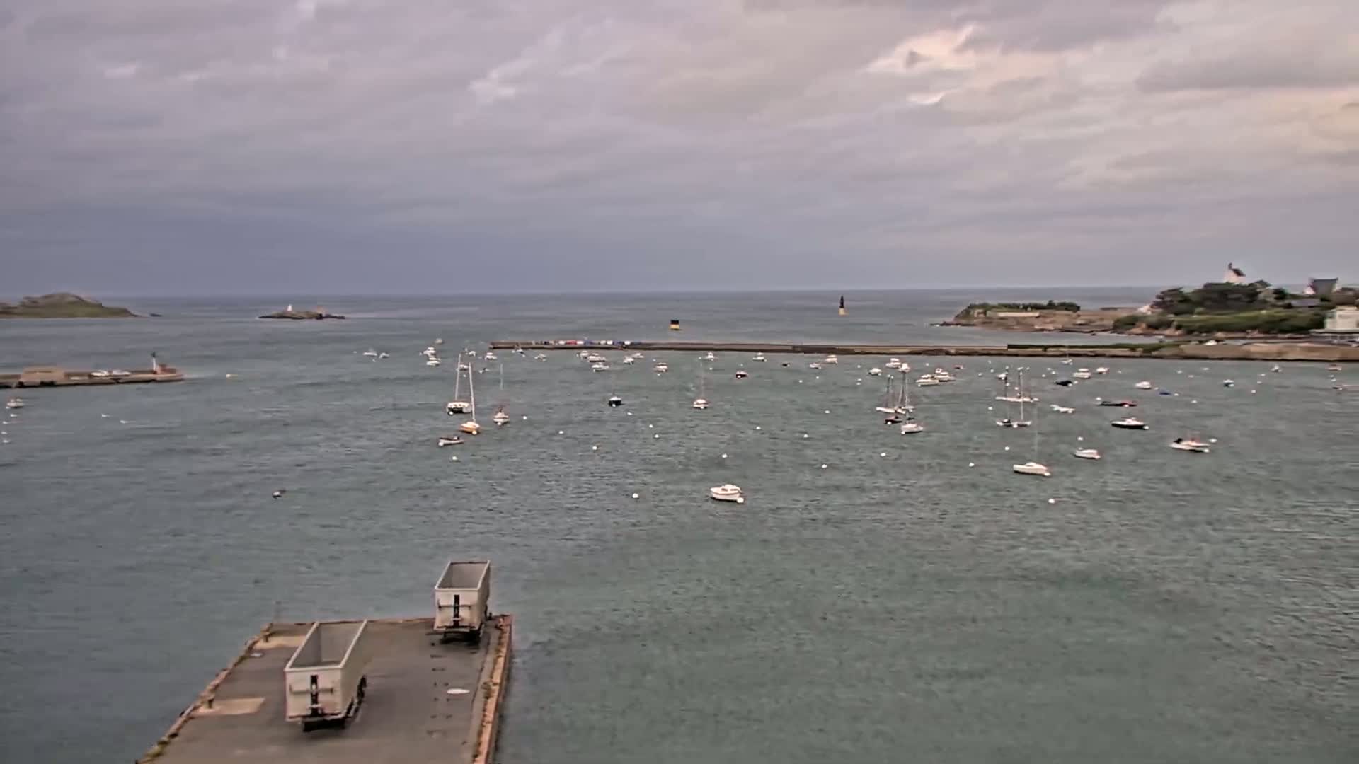 Under an overcast sky, numerous small boats are moored in a calm harbor, with larger landmasses visible in the distance.