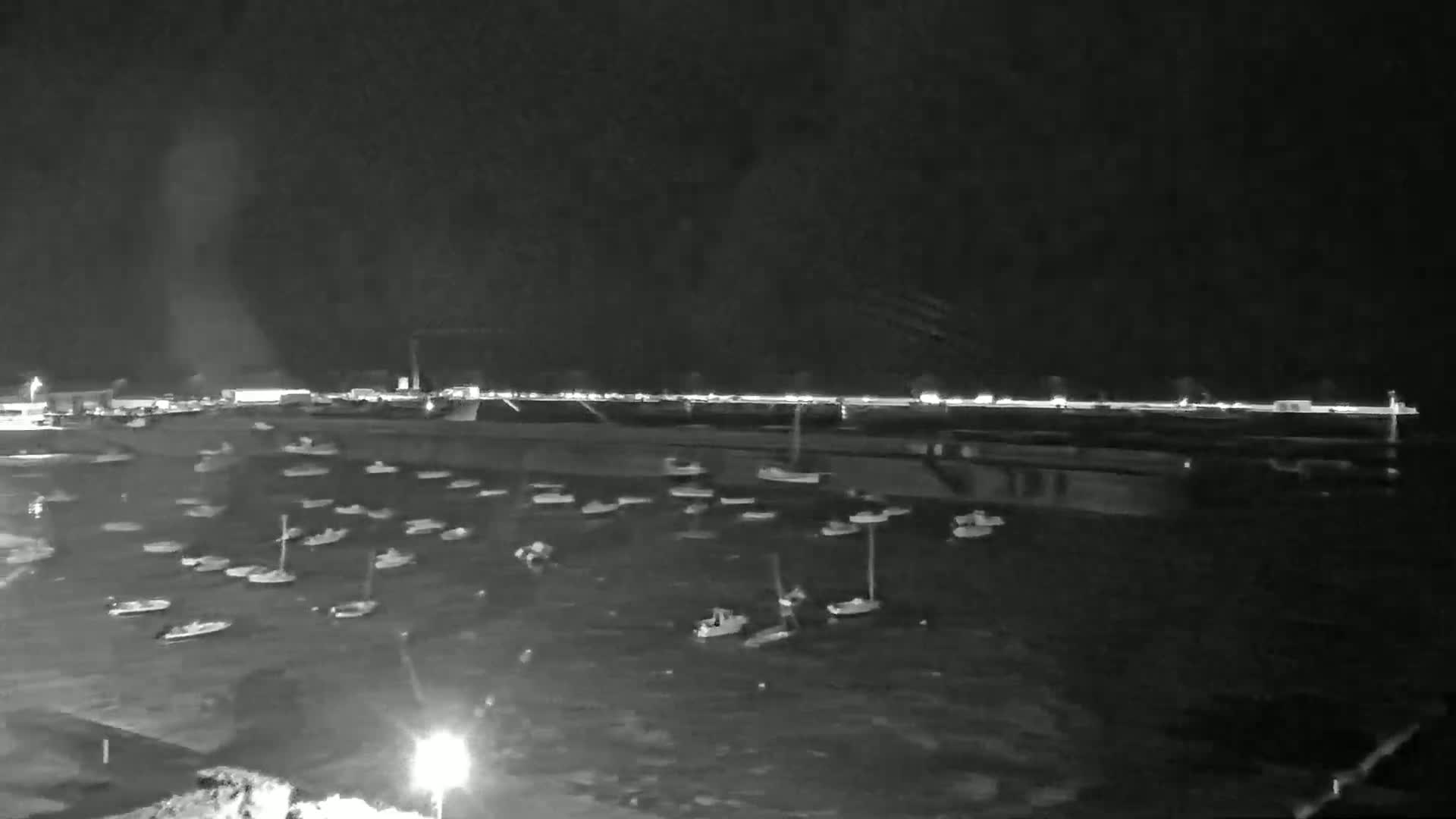 A nighttime, low-light view shows numerous small boats moored in calm water near a brightly lit shoreline.