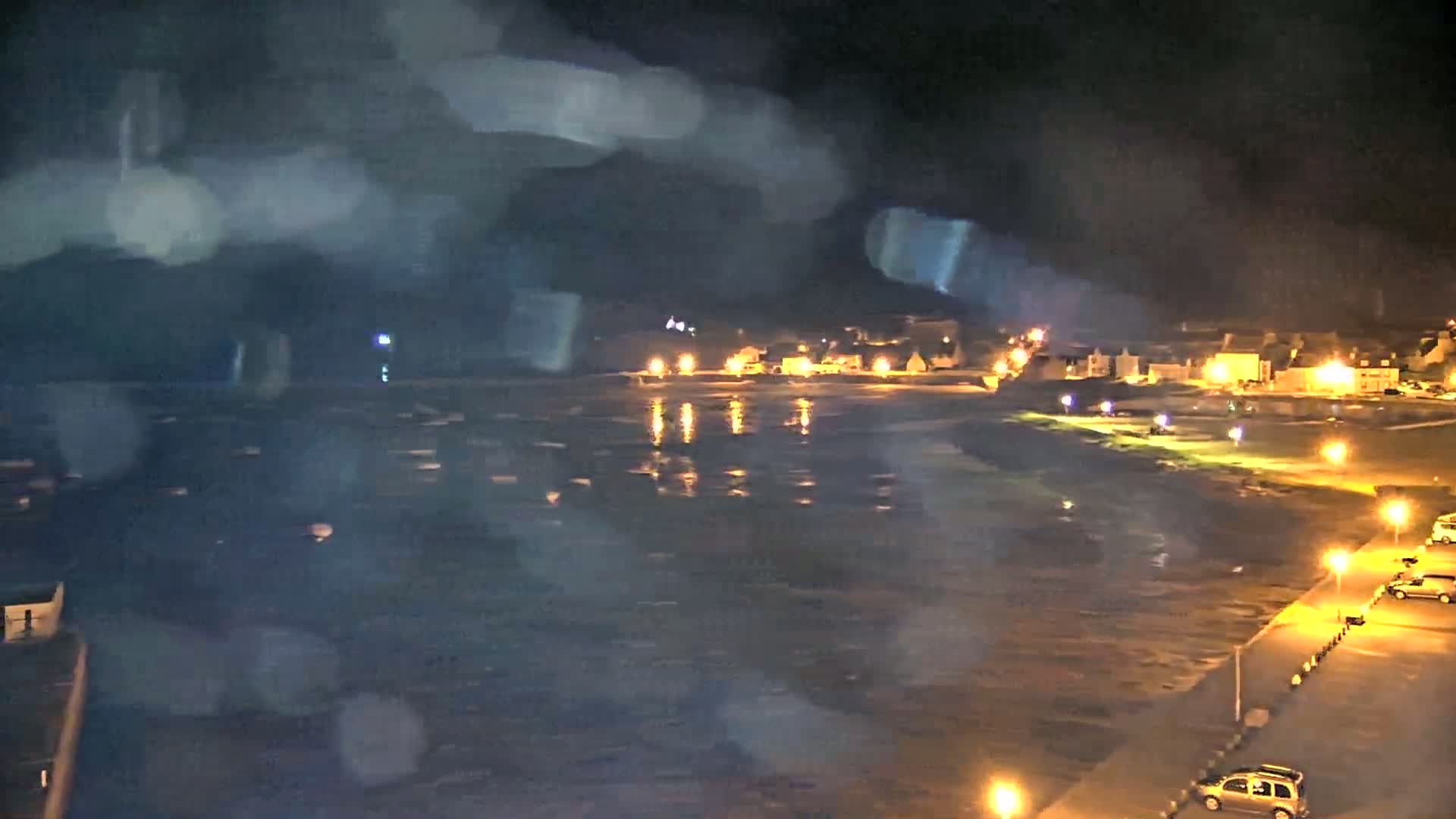 A nighttime view of a rain-soaked coastal town, with its lights reflecting on the dark water.