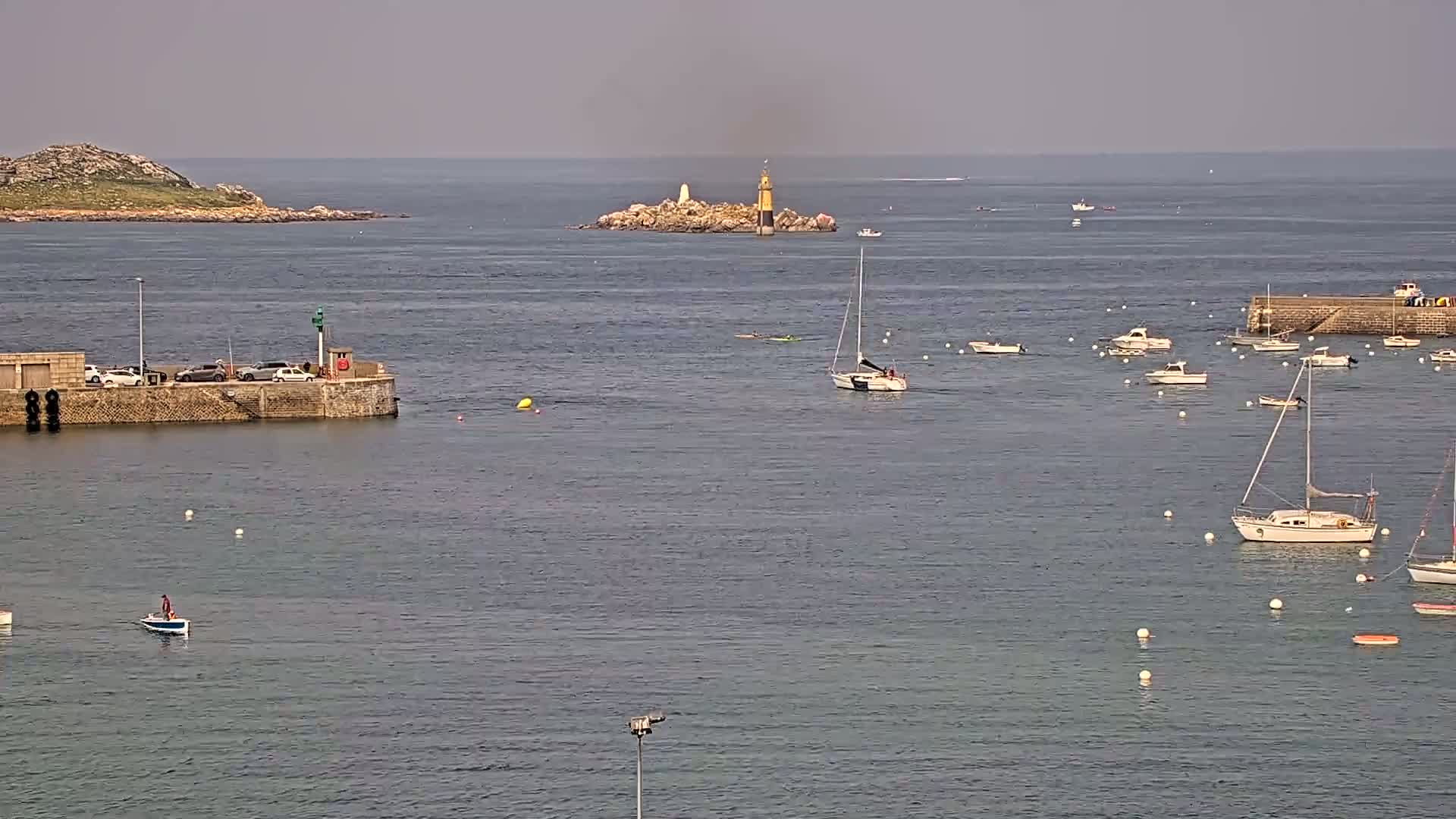 A calm harbor scene shows numerous small boats and sailboats anchored near a stone pier and island with a lighthouse under an overcast sky.