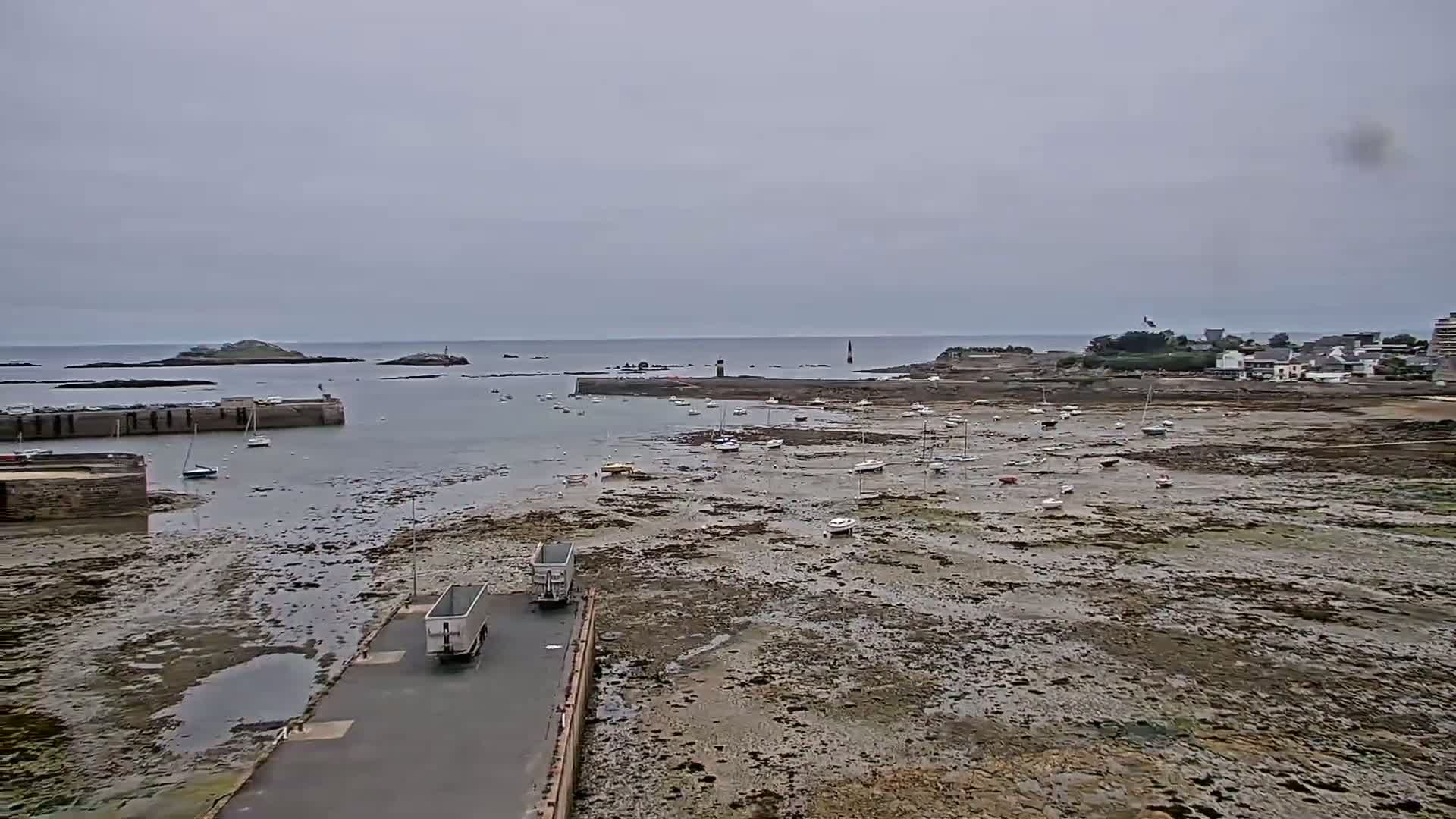 Under an overcast sky, a harbor reveals numerous small boats resting on exposed mudflats at low tide, with two trucks parked near a paved road leading to the water.