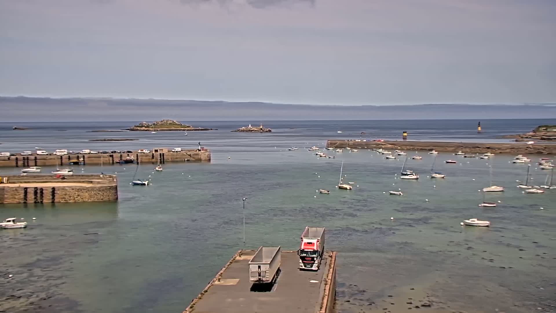A harbor filled with numerous sailboats and a few motorboats under a mostly cloudy sky, with two semi-trailer trucks parked on a pier, and a small island visible in the distance.