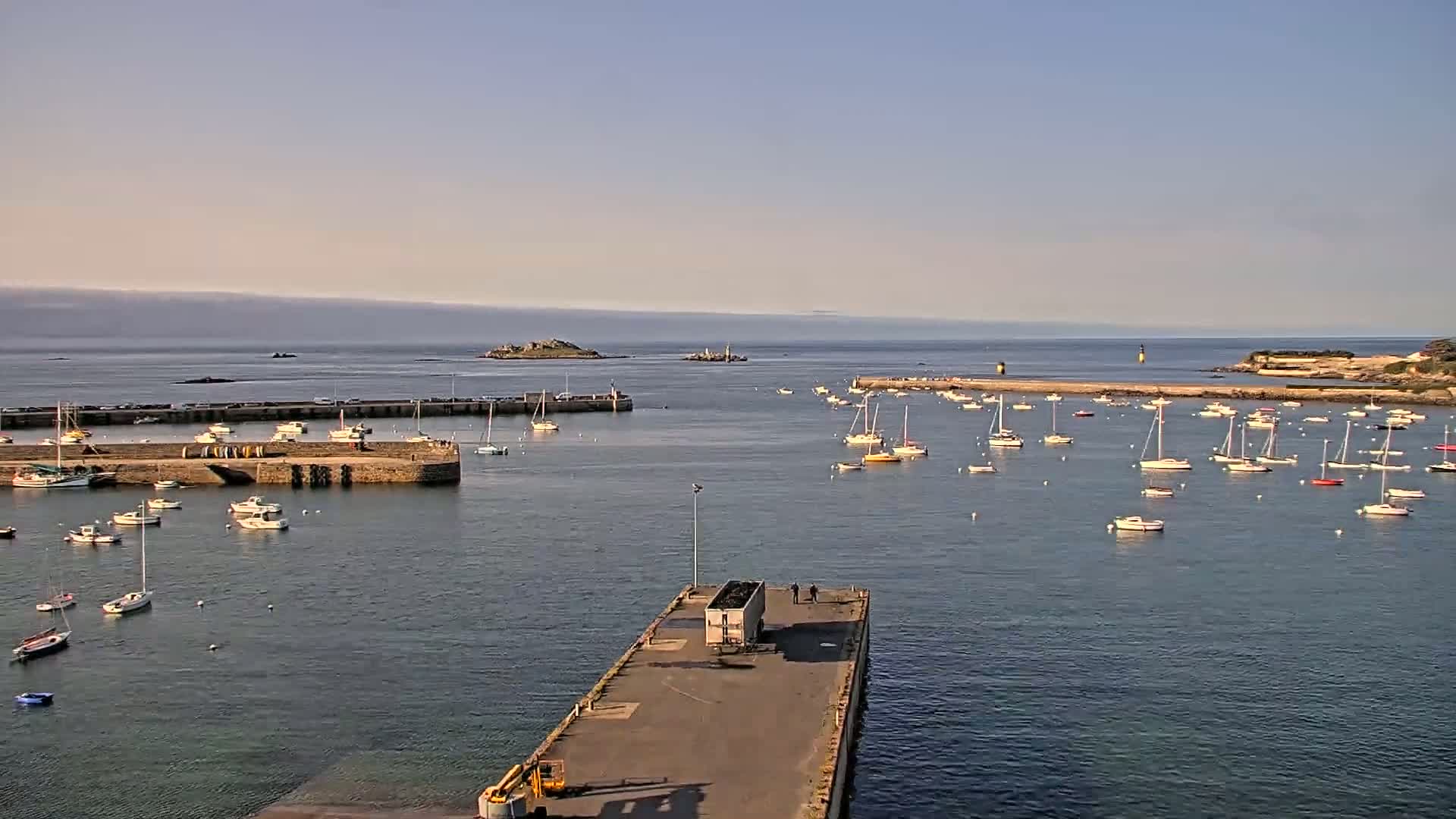 A calm harbor filled with numerous sailboats and small motorboats, under a clear sky, with an island visible in the distance.