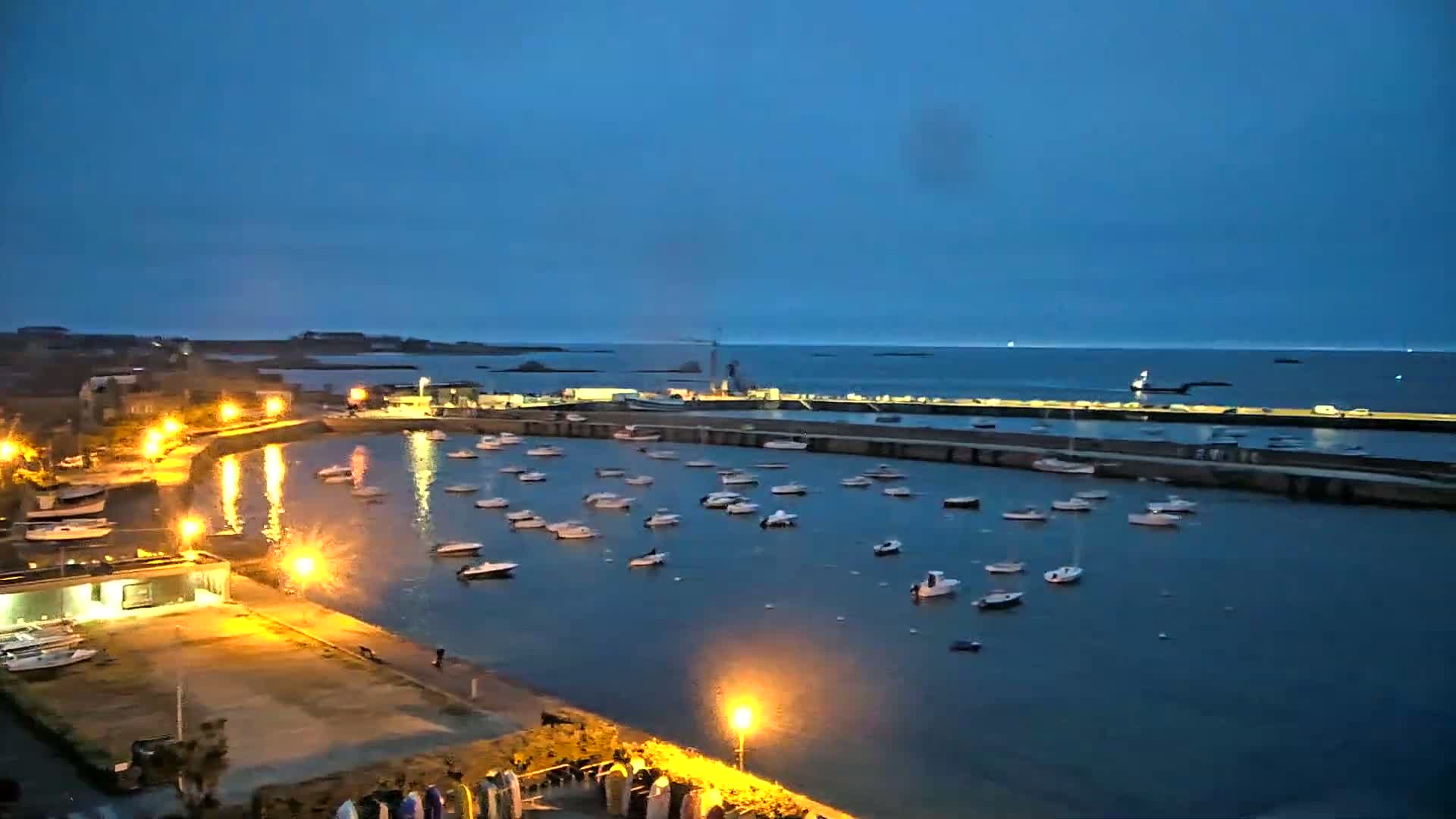 A harbor filled with numerous small boats is illuminated at night under a clear, dark blue twilight sky.