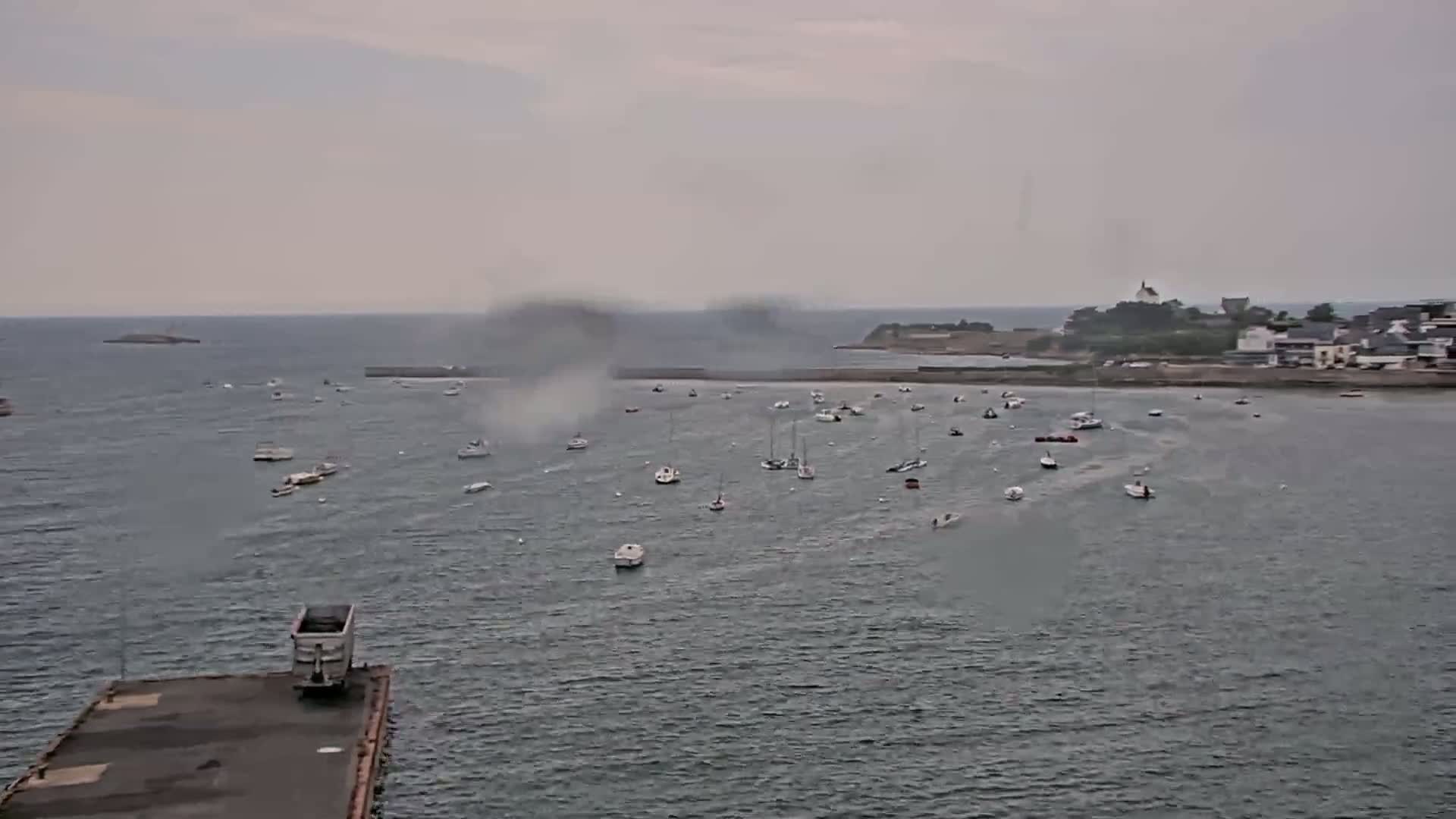 Many small boats are in a calm, gray harbor under an overcast sky, with a landmass and buildings visible in the distance.