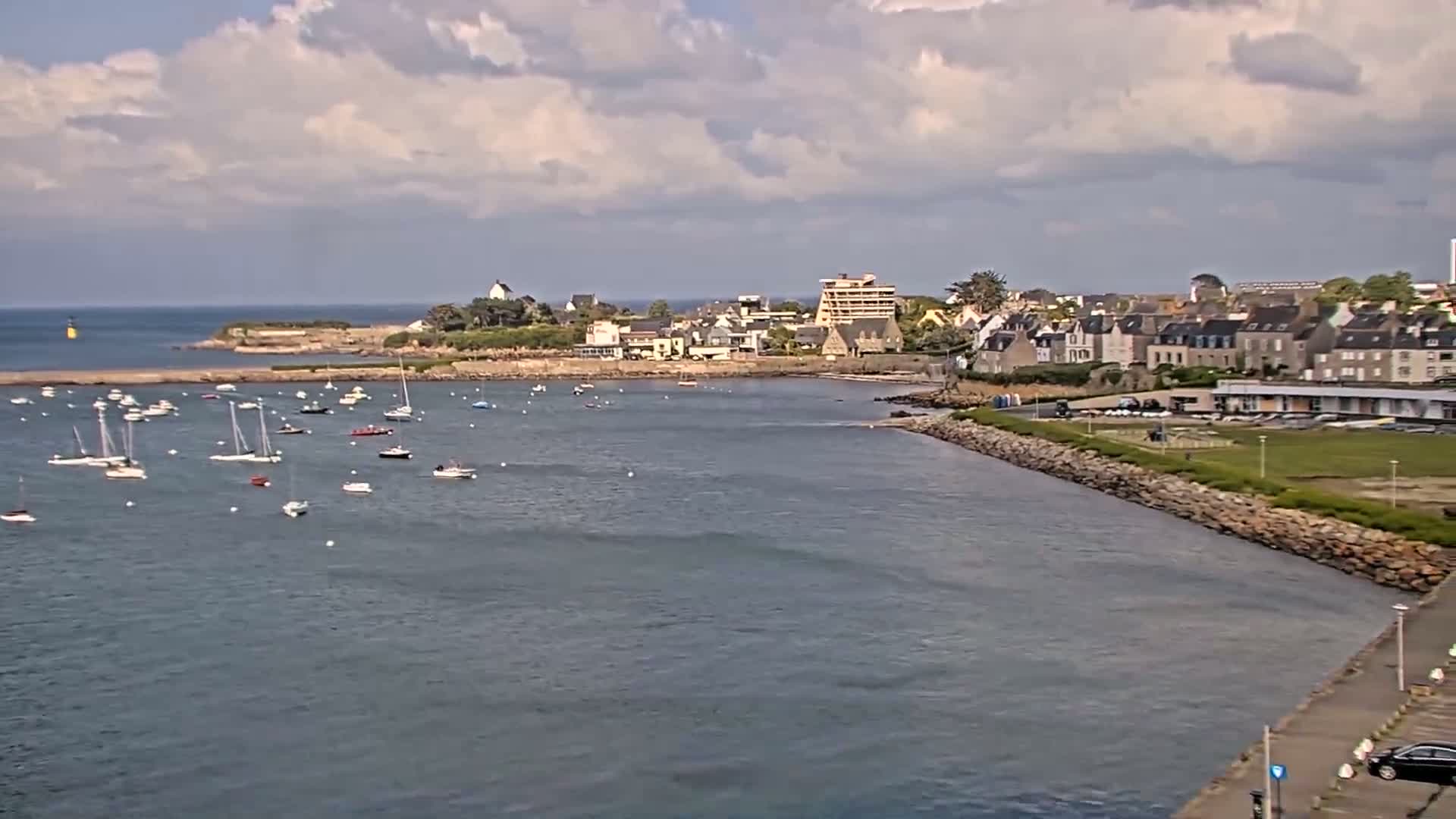 A partly cloudy day shows a calm harbor filled with sailboats near a town situated on a rocky coast.
