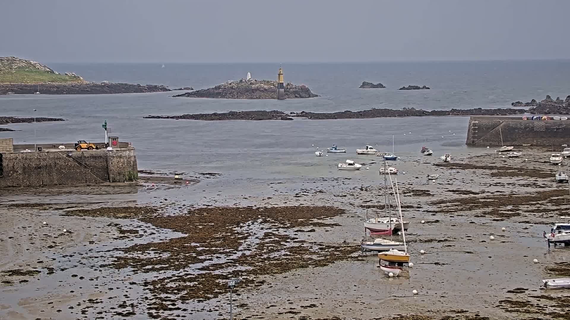 A harbor with several boats sits exposed at low tide under an overcast sky, with a small island and lighthouse visible in the distance.