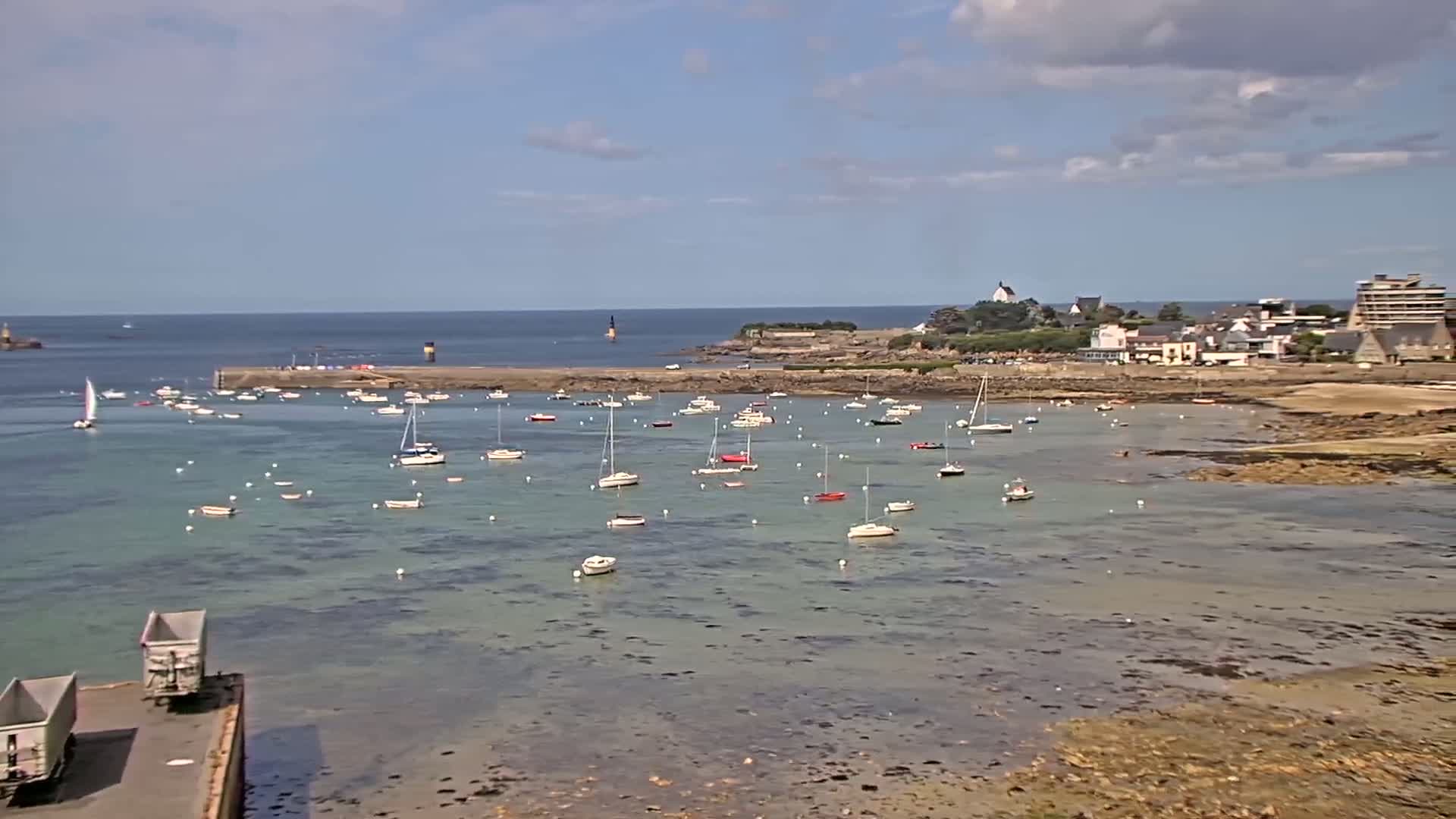 Many sailboats are moored in a calm, shallow harbor on a partly sunny day, with a small town visible in the background.