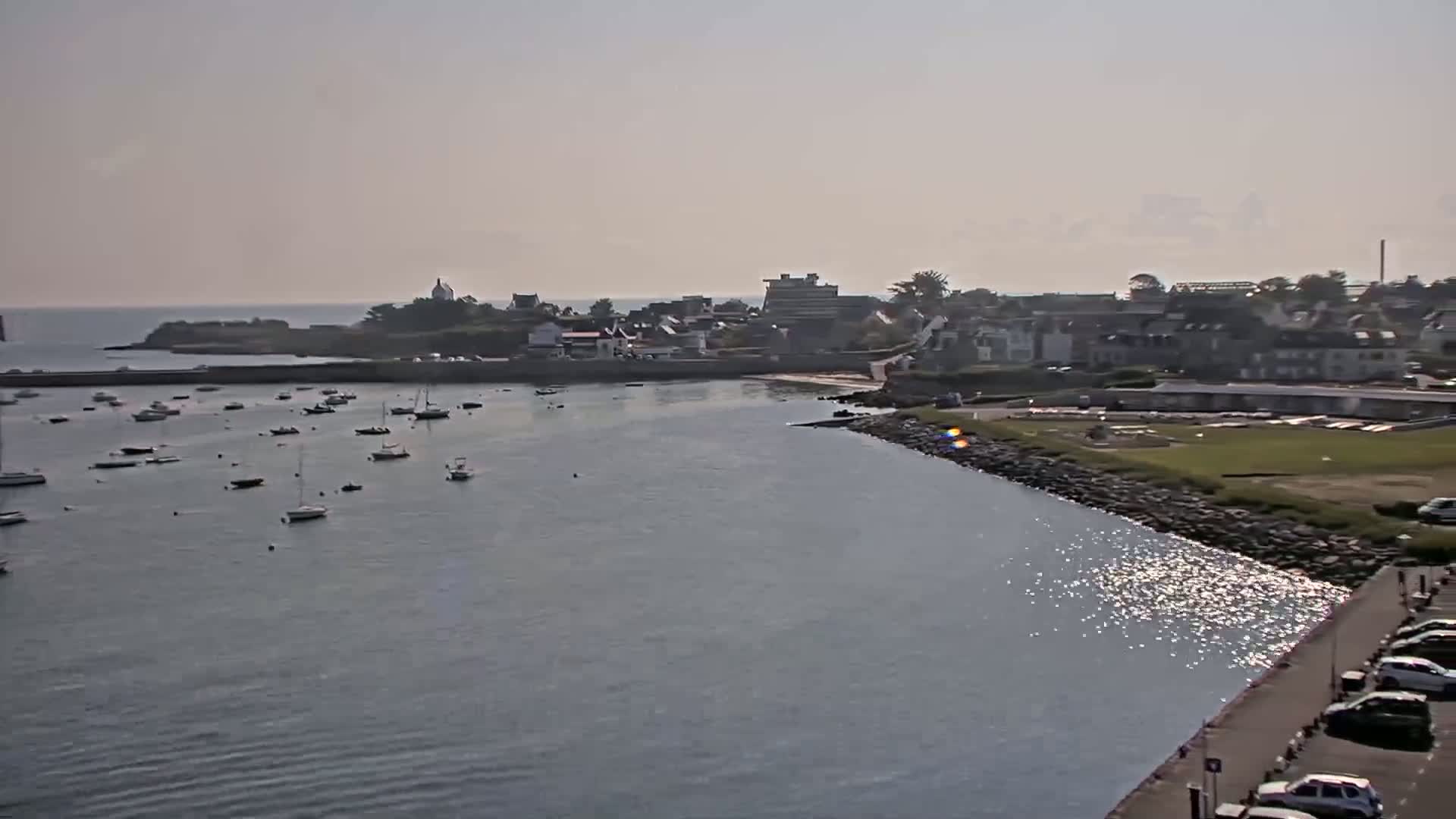 A calm harbor filled with numerous small boats is nestled beside a coastal town under a hazy, sunny sky.