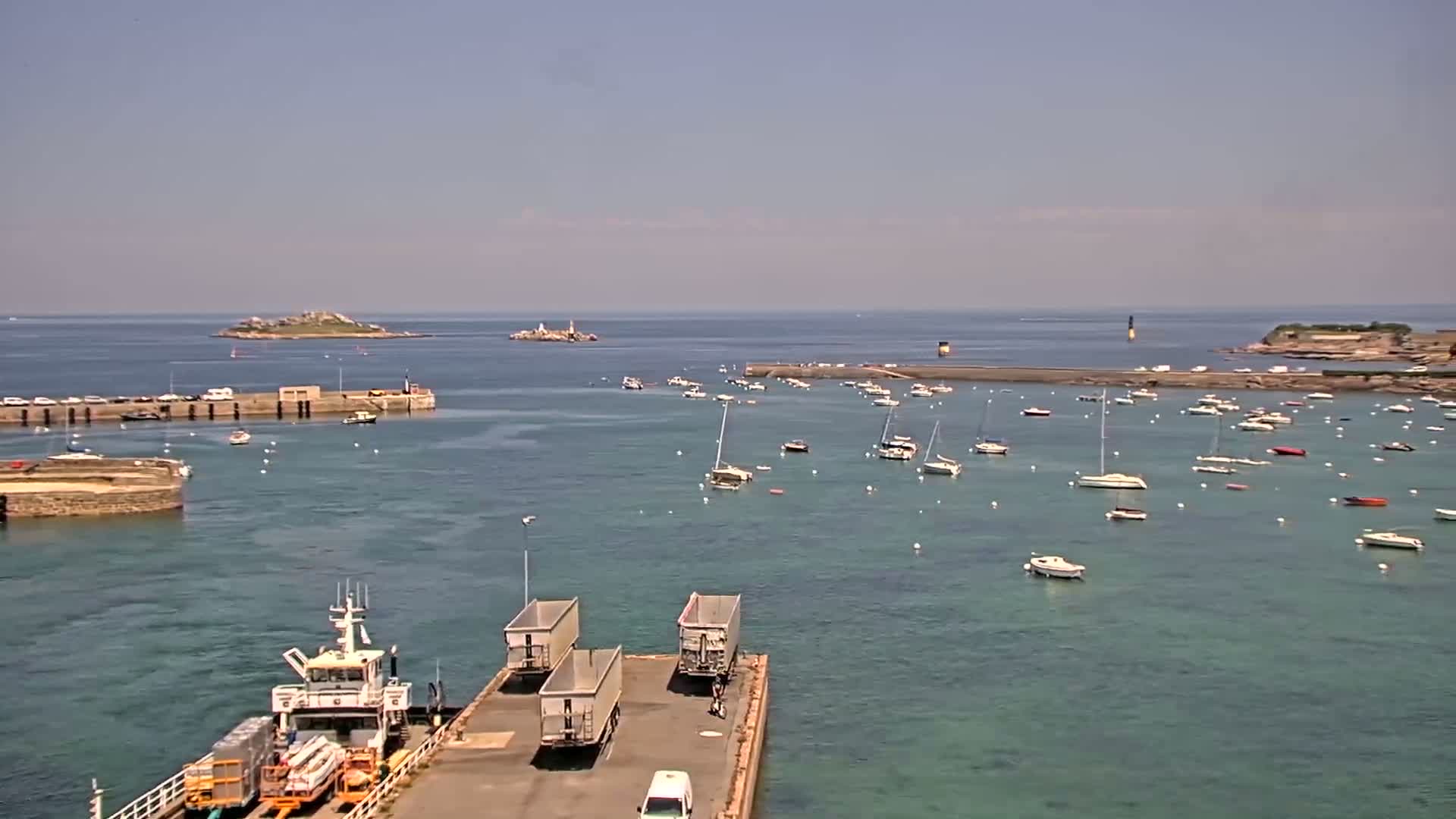 A harbor filled with sailboats under a clear sky shows a ferry docked with cargo containers and two small islands in the distance.