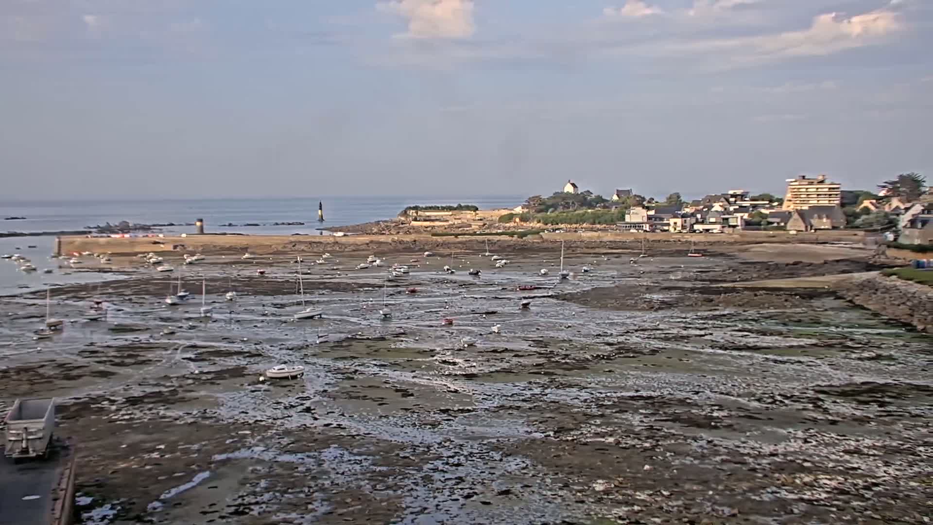 A mostly sunny day shows a harbor with numerous small sailboats resting on exposed mudflats at low tide, with a small town and distant ocean in the background.