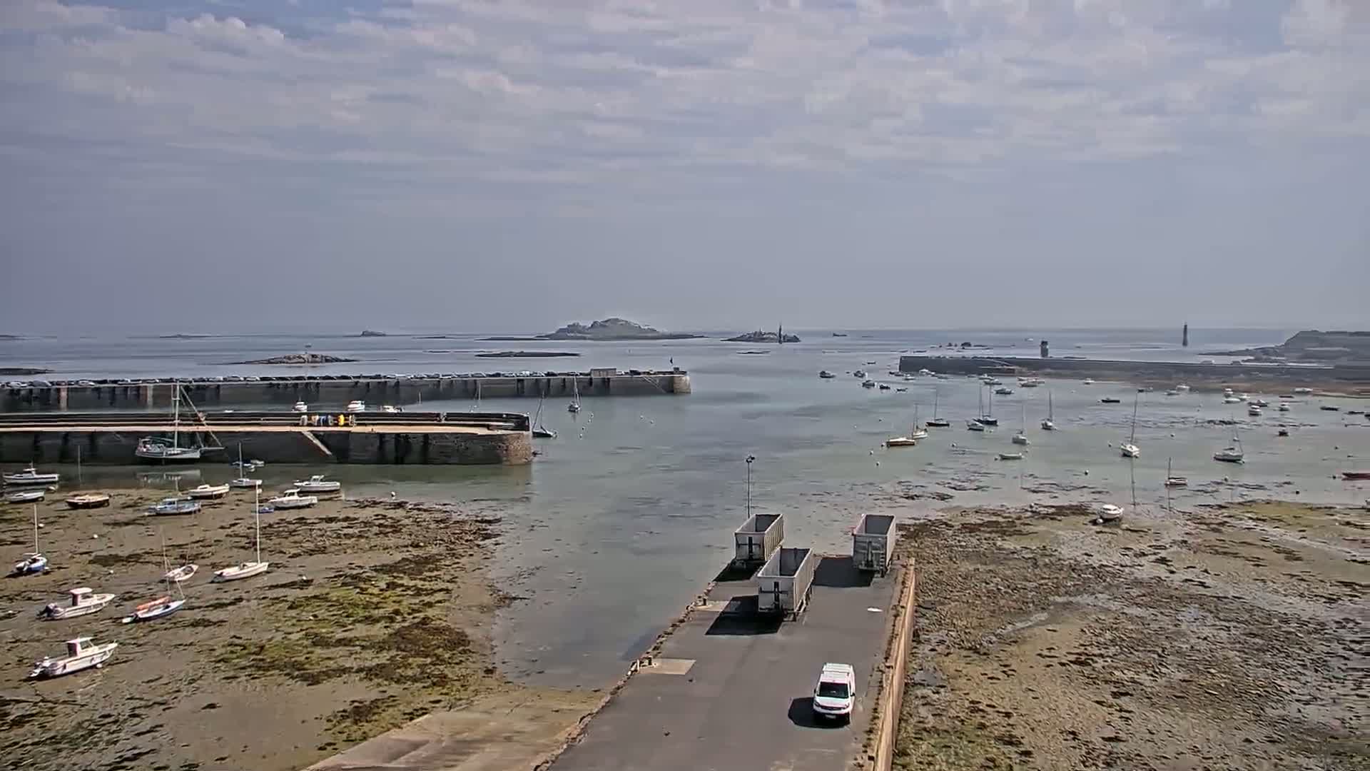A harbor with numerous boats at low tide under a partly cloudy sky, with several semi-trucks and a van parked on a road near the water.