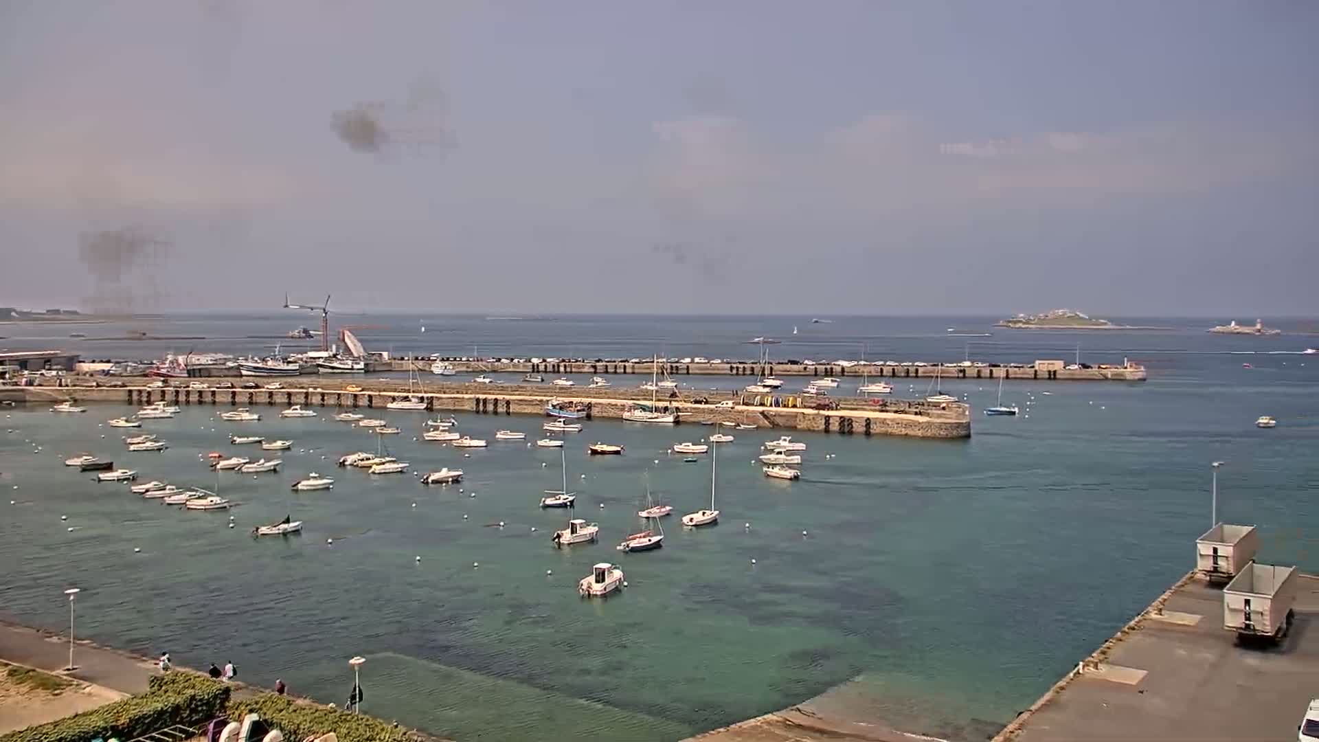 A harbor filled with numerous small boats under a hazy, sunny sky, with a larger body of water and a distant island visible in the background.