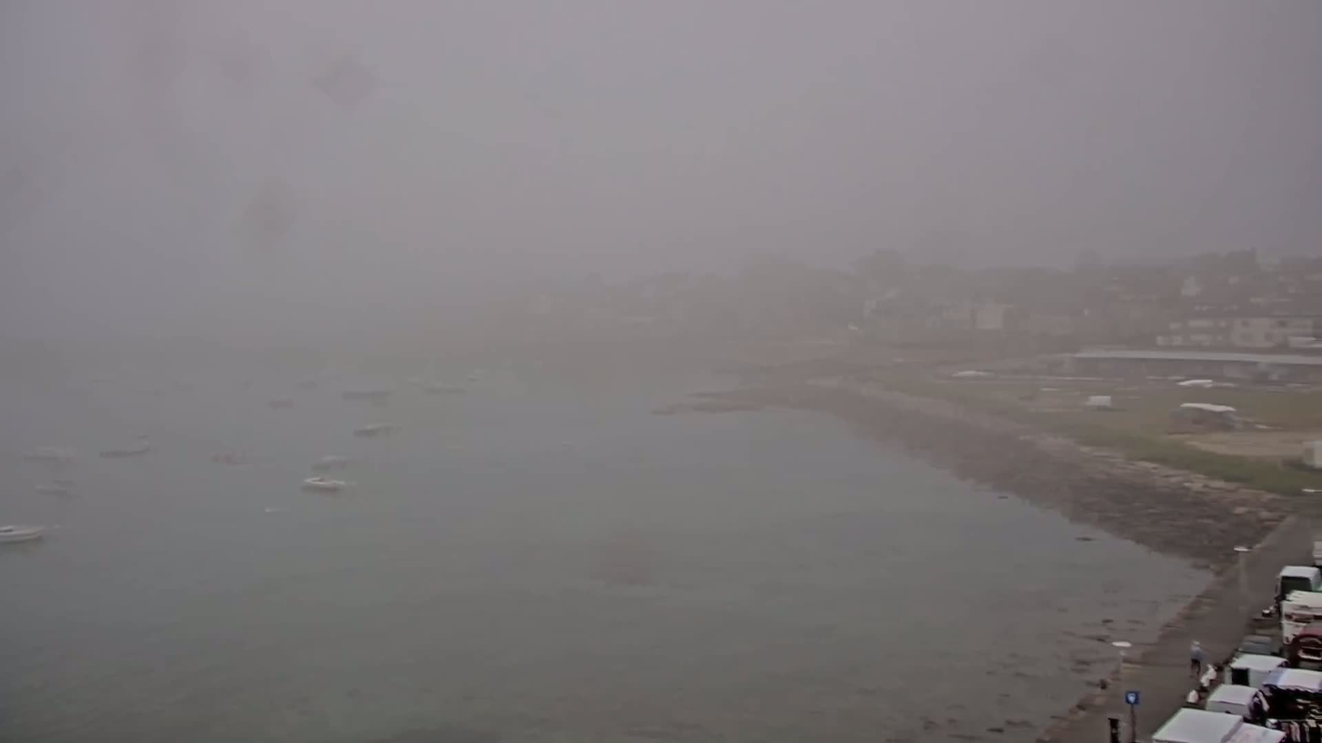 A hazy, foggy coastal scene shows a calm body of water with several small boats, a shoreline with buildings and a paved area, and some vehicles parked near the water.