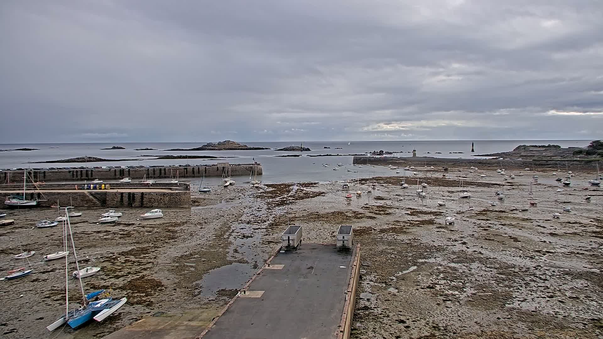 A harbor filled with numerous boats sits exposed at low tide under an overcast sky, with small islands visible in the distance.