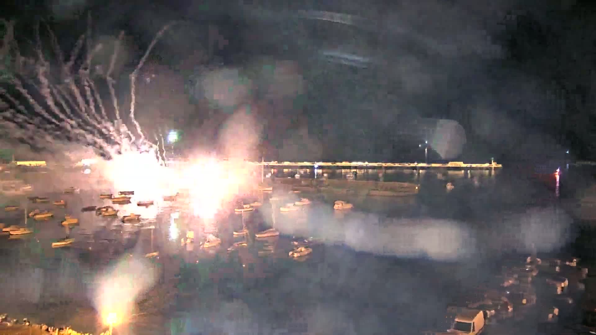 A nighttime fireworks display over a harbor filled with numerous small boats, with smoke and some light rain obscuring the view.