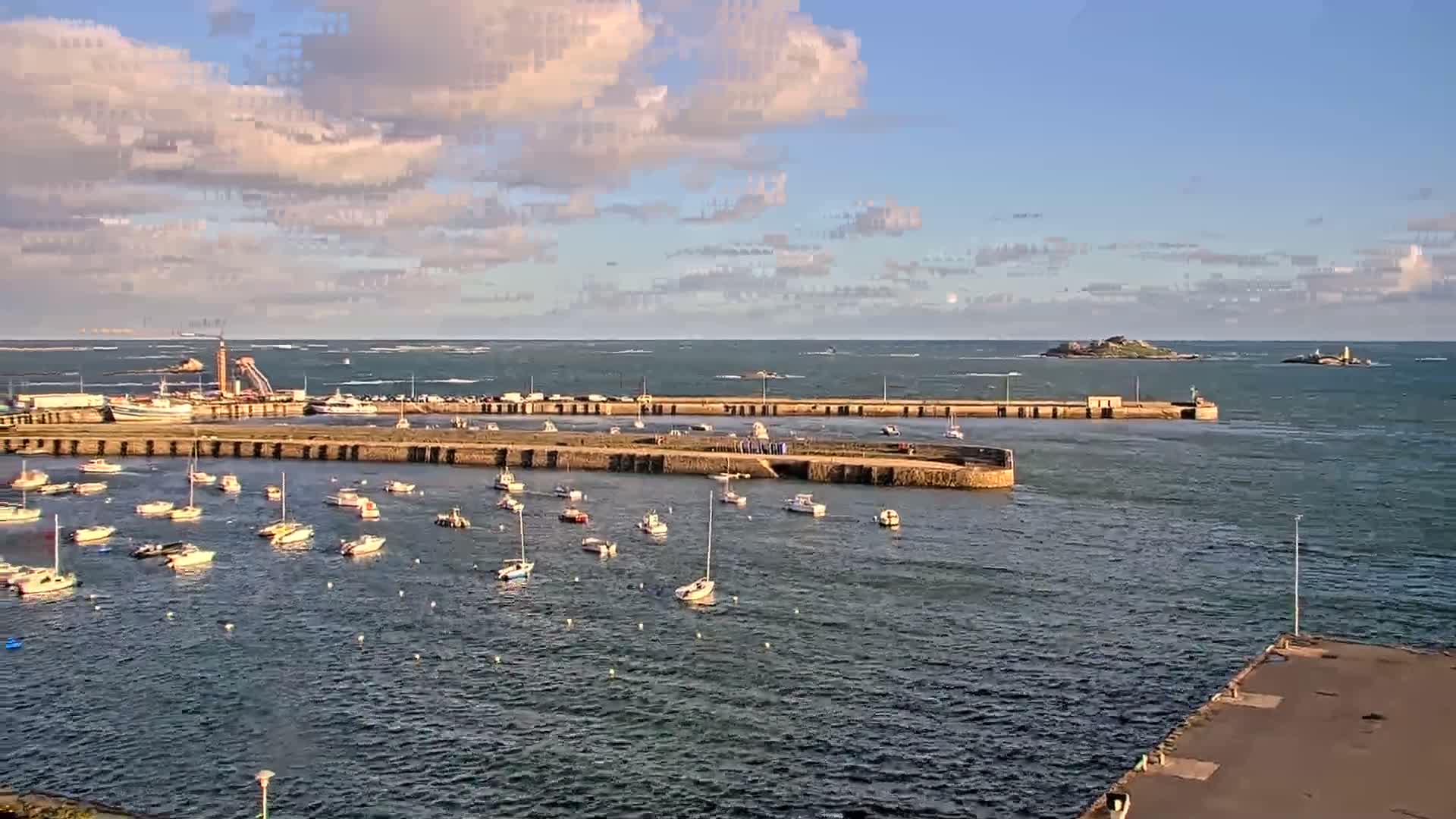 The image shows a sunny day at a harbor filled with numerous moored boats, featuring long stone breakwaters, a distant industrial port with larger vessels, and two islands on the horizon under a bright blue sky with scattered clouds.