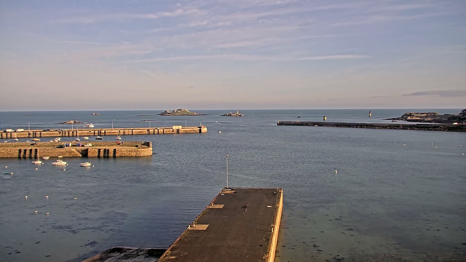 The image shows a sunny day at a harbor filled with numerous moored boats, featuring long stone breakwaters, a distant industrial port with larger vessels, and two islands on the horizon under a bright blue sky with scattered clouds.