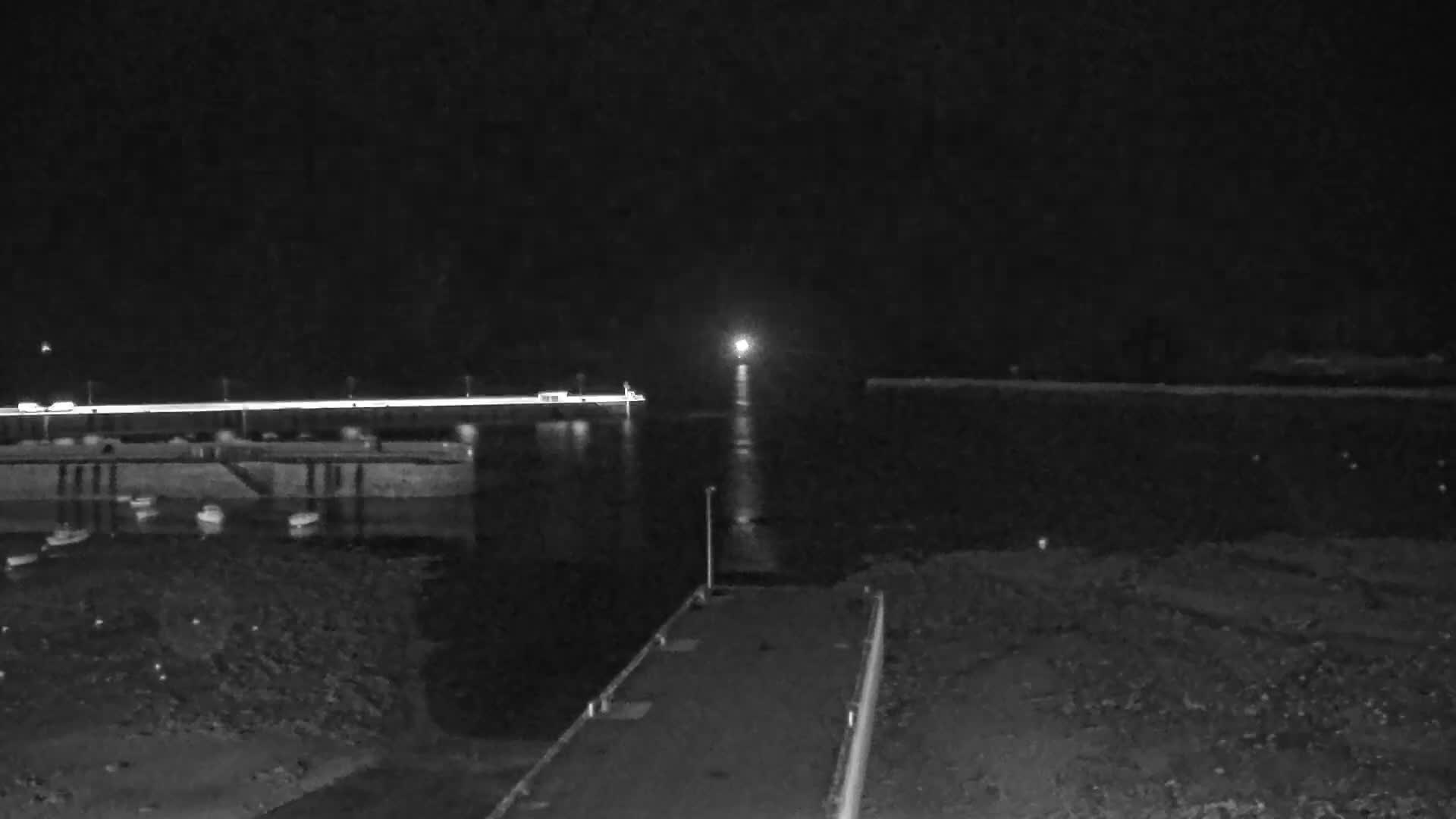 The image shows a sunny day at a harbor filled with numerous moored boats, featuring long stone breakwaters, a distant industrial port with larger vessels, and two islands on the horizon under a bright blue sky with scattered clouds.