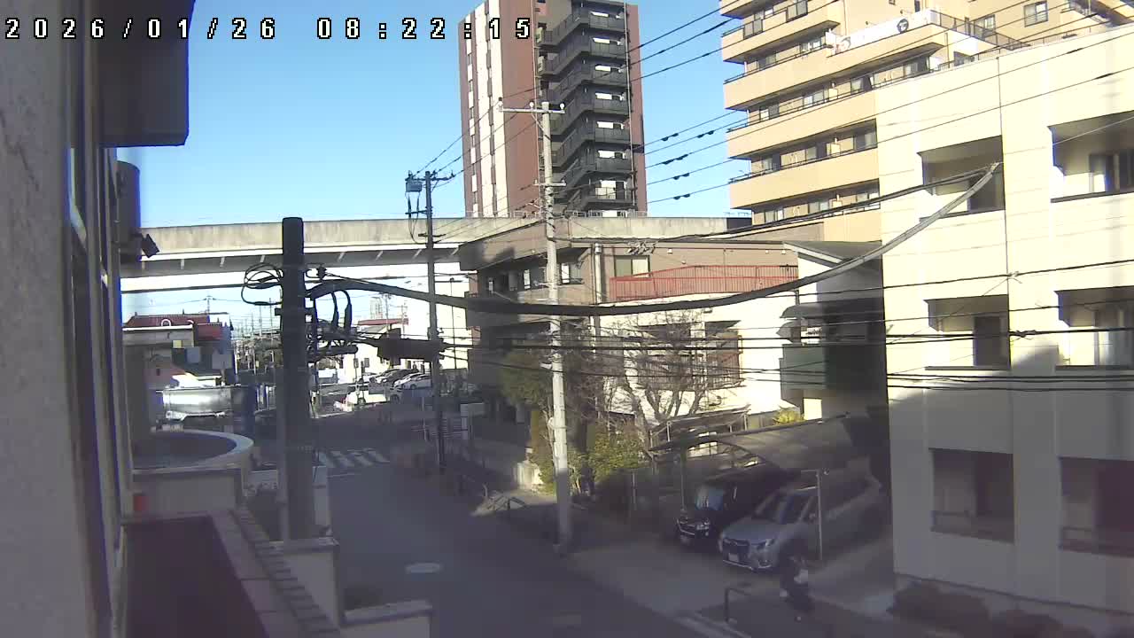 From an elevated perspective, a residential street with diverse buildings, parked cars, prominent utility poles and wires, and an elevated concrete overpass unfolds under a clear and sunny sky.