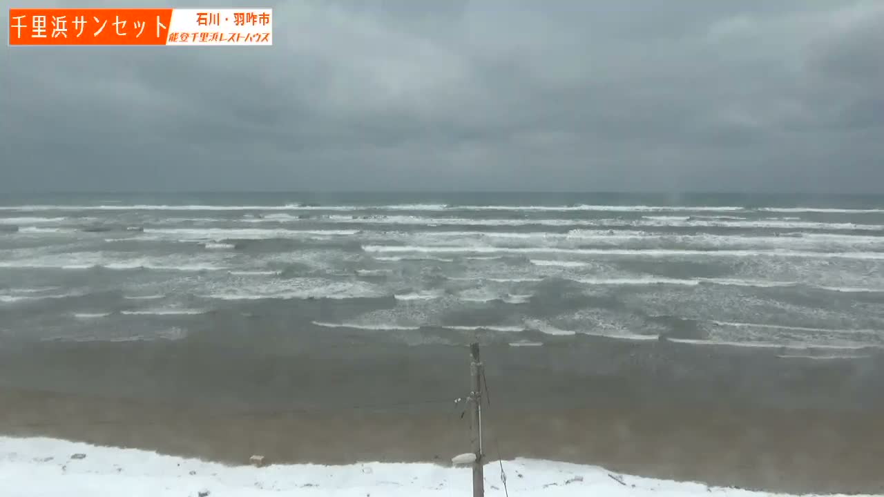A wintry coastline features a rough, grey ocean with white-capped waves meeting a snow-dusted sandy beach under a heavily overcast and gloomy sky.