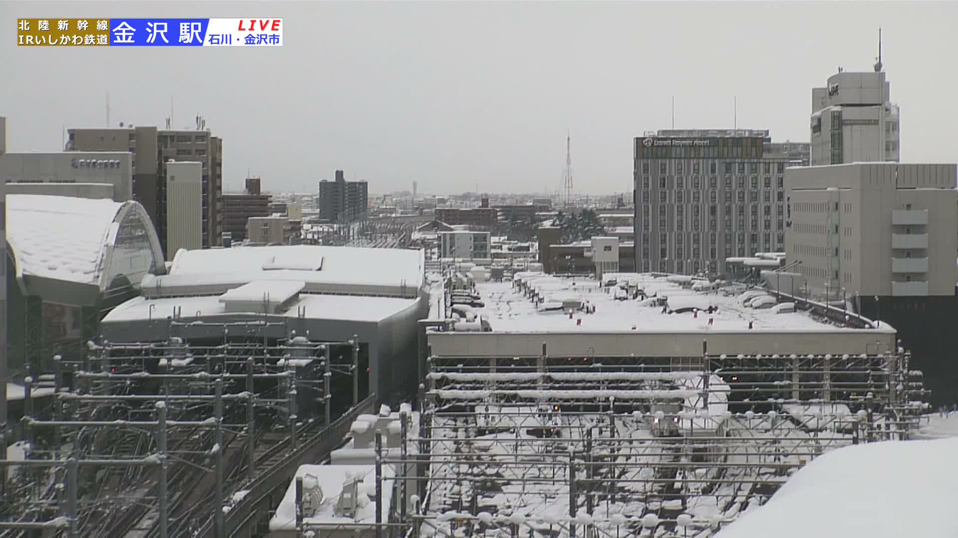 A dense urban landscape is visible under an overcast sky, with rooftops, train tracks, and extensive overhead wiring structures heavily blanketed in fresh snow.