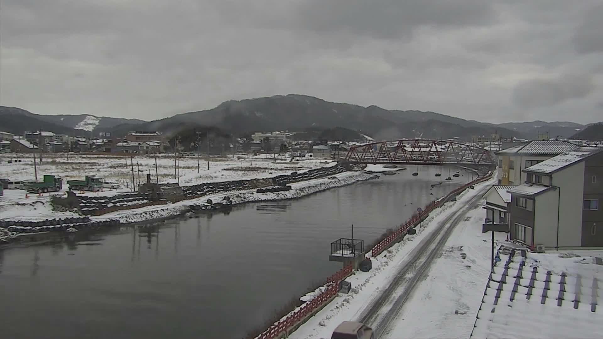 Under an overcast sky, a wide river flows through a snow-covered town featuring a distinctive red truss bridge and distant mountains.
