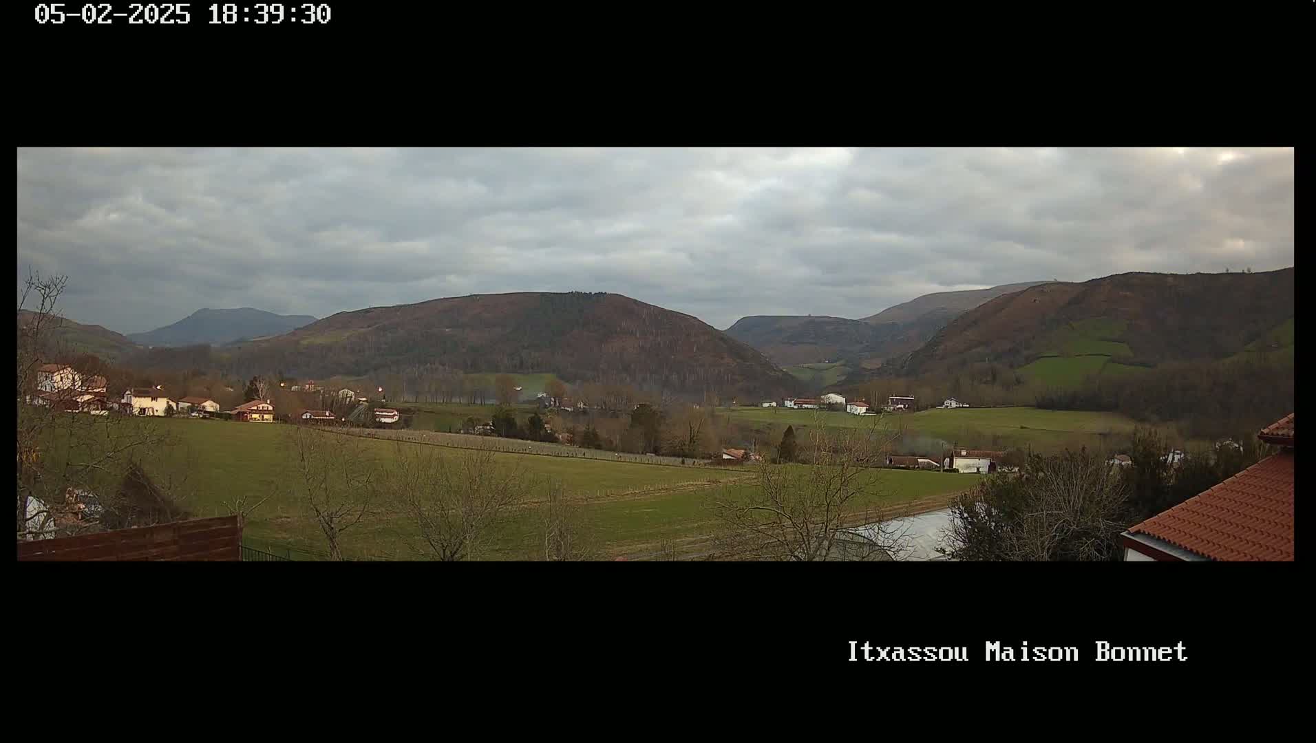 A rural valley landscape under a cloudy sky shows several houses nestled among green fields and rolling hills.